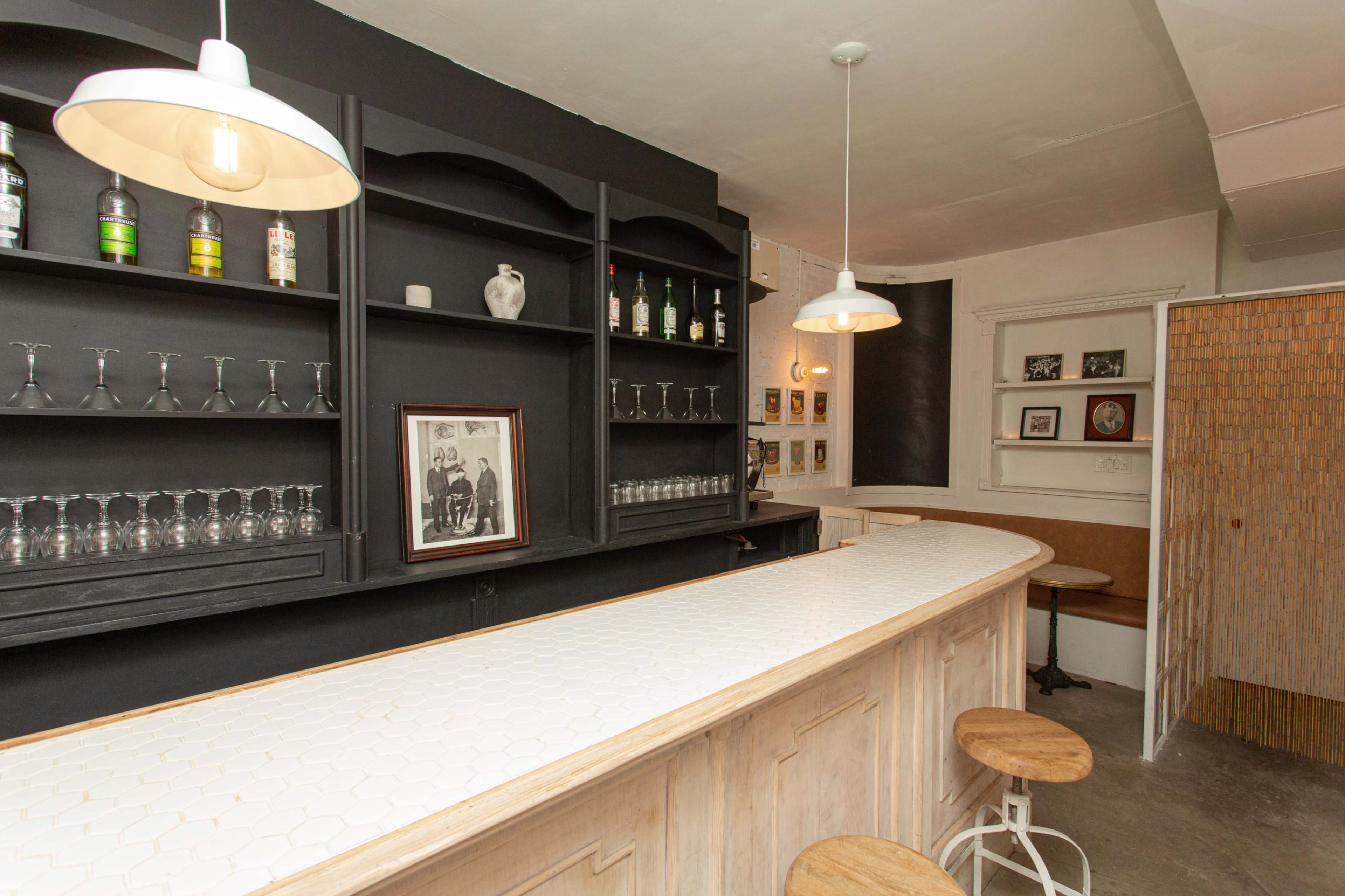 The image shows a bar with a white hexagonal-tiled counter, black shelves displaying bottles and glassware, and wooden stools at the front.