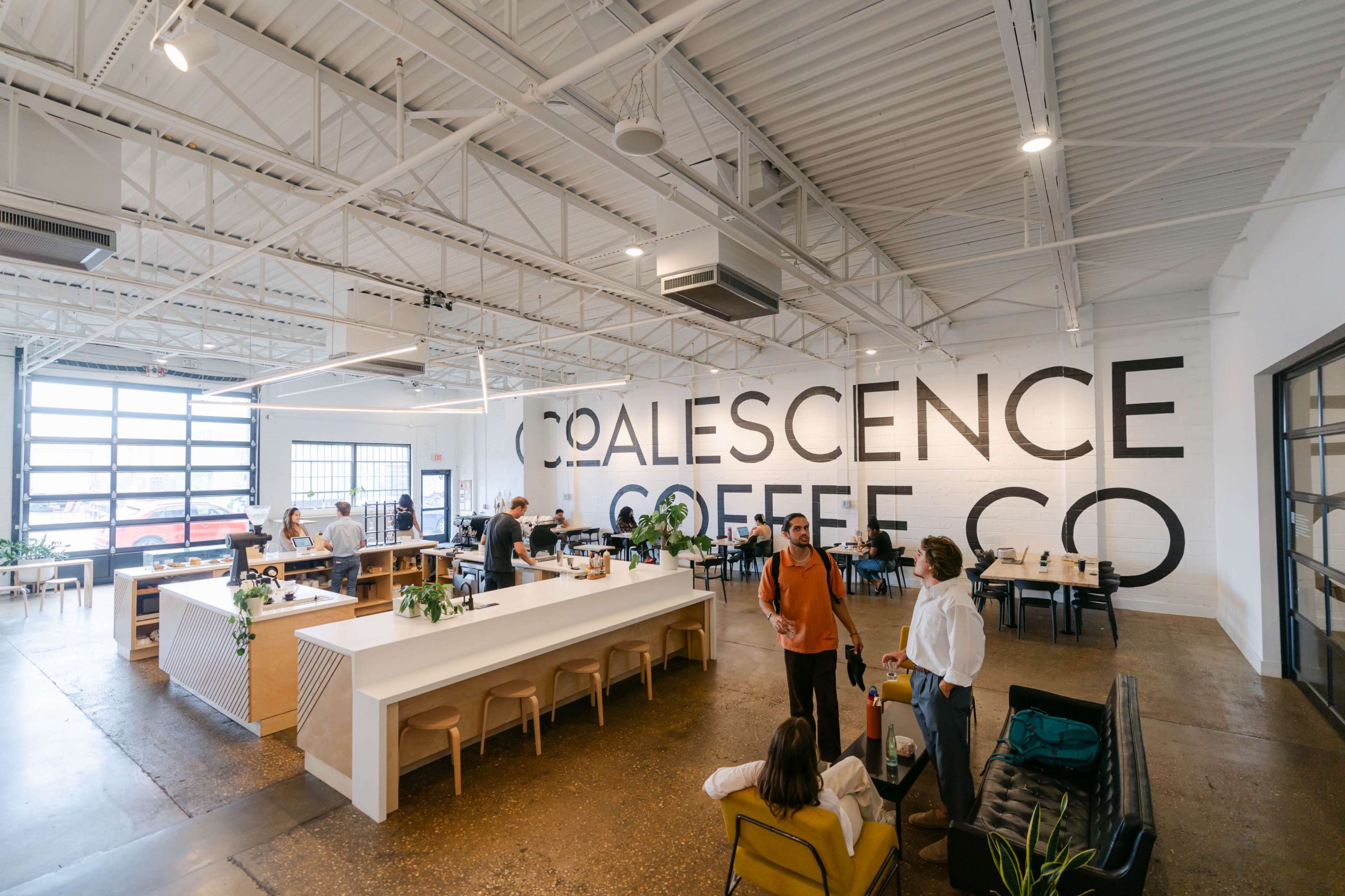 A spacious coffee shop interior with a large "Coalescence Coffee Co" mural on the wall, featuring a central counter, seating areas, and customers interacting.