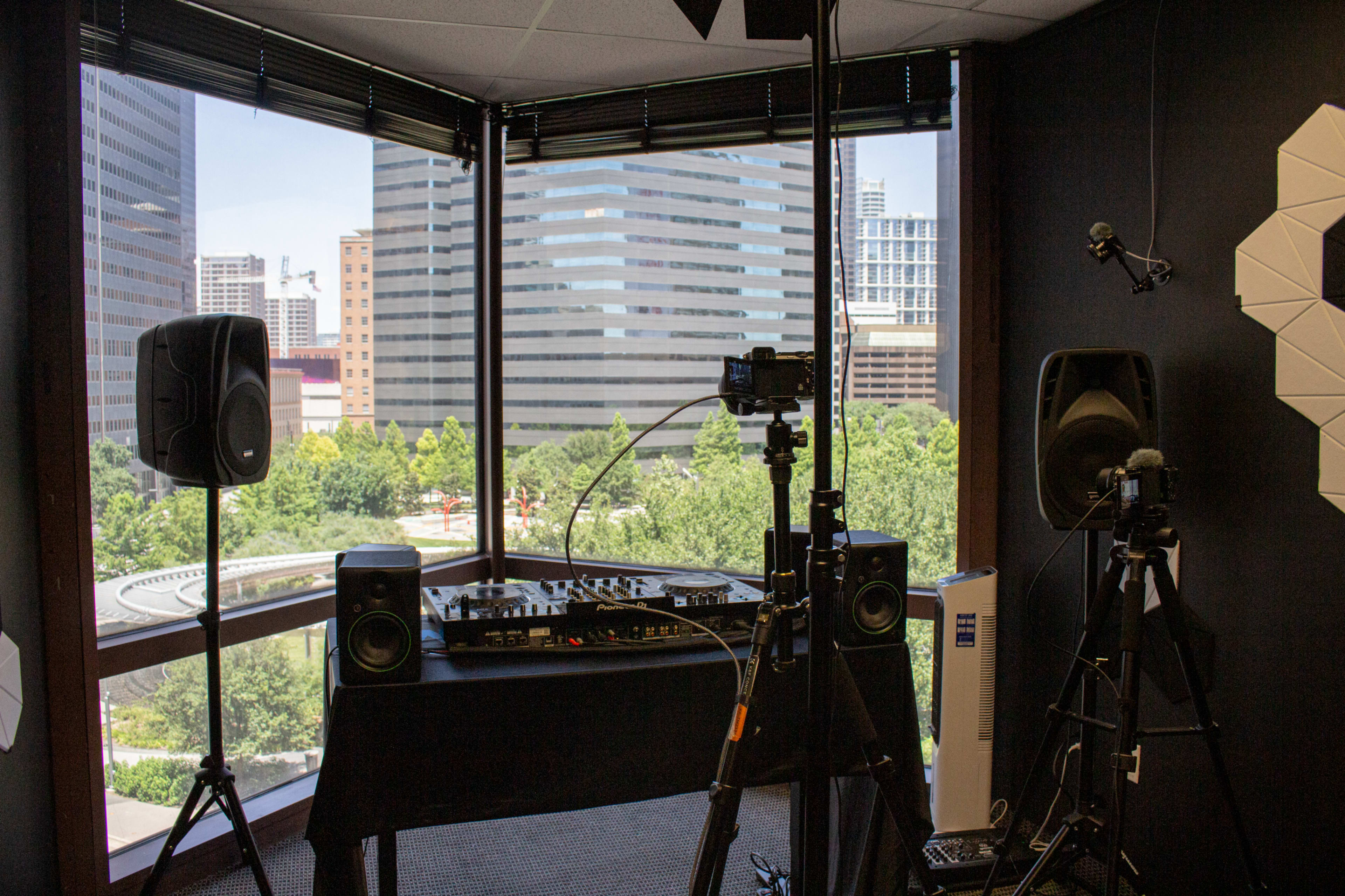 The image shows a modern recording studio setup with a DJ mixer and speakers positioned by a large window overlooking a cityscape and greenery.