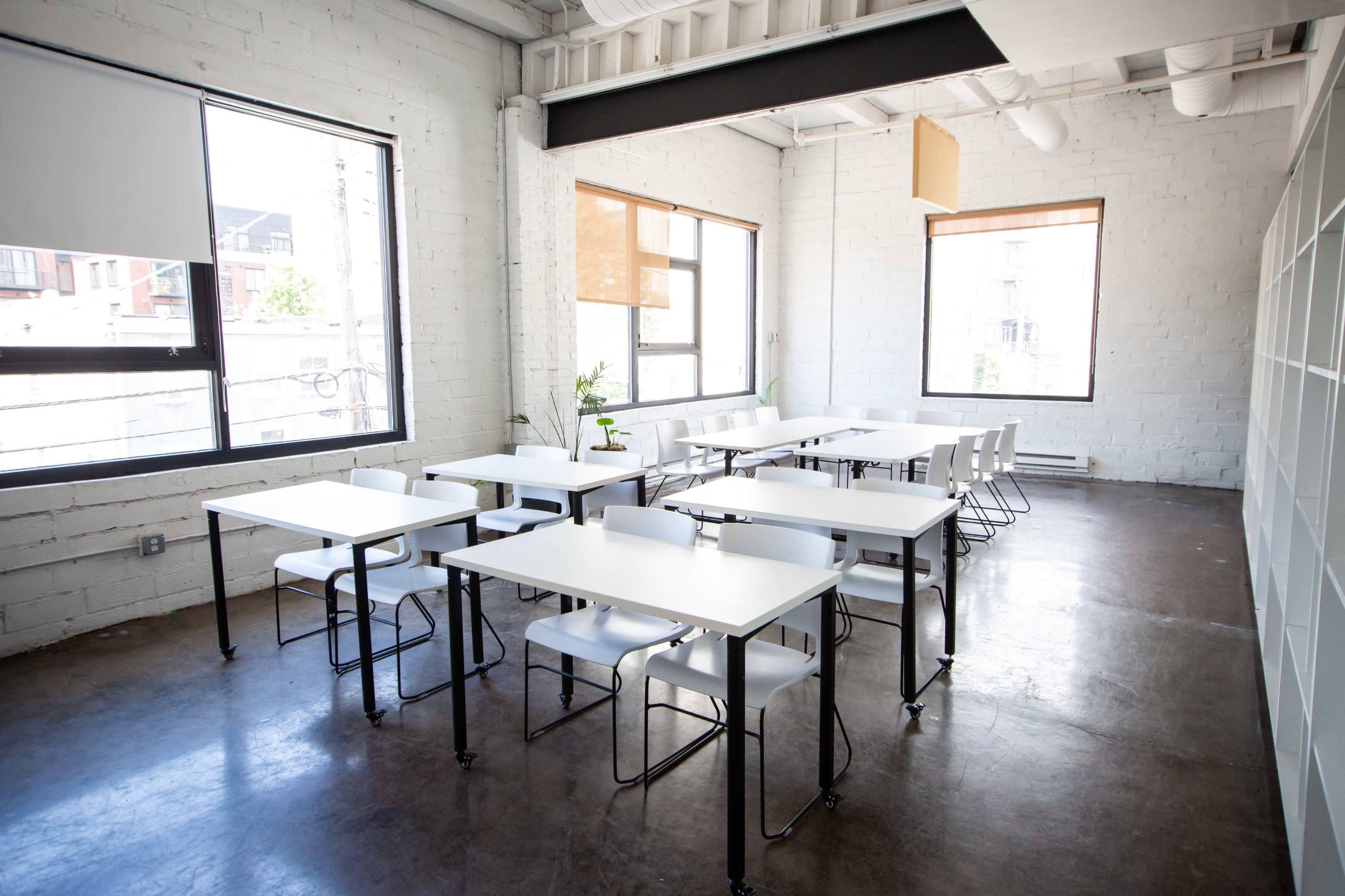 The image shows a modern classroom with several white tables and chairs arranged neatly, large windows allowing natural light to enter, and a minimalist design.