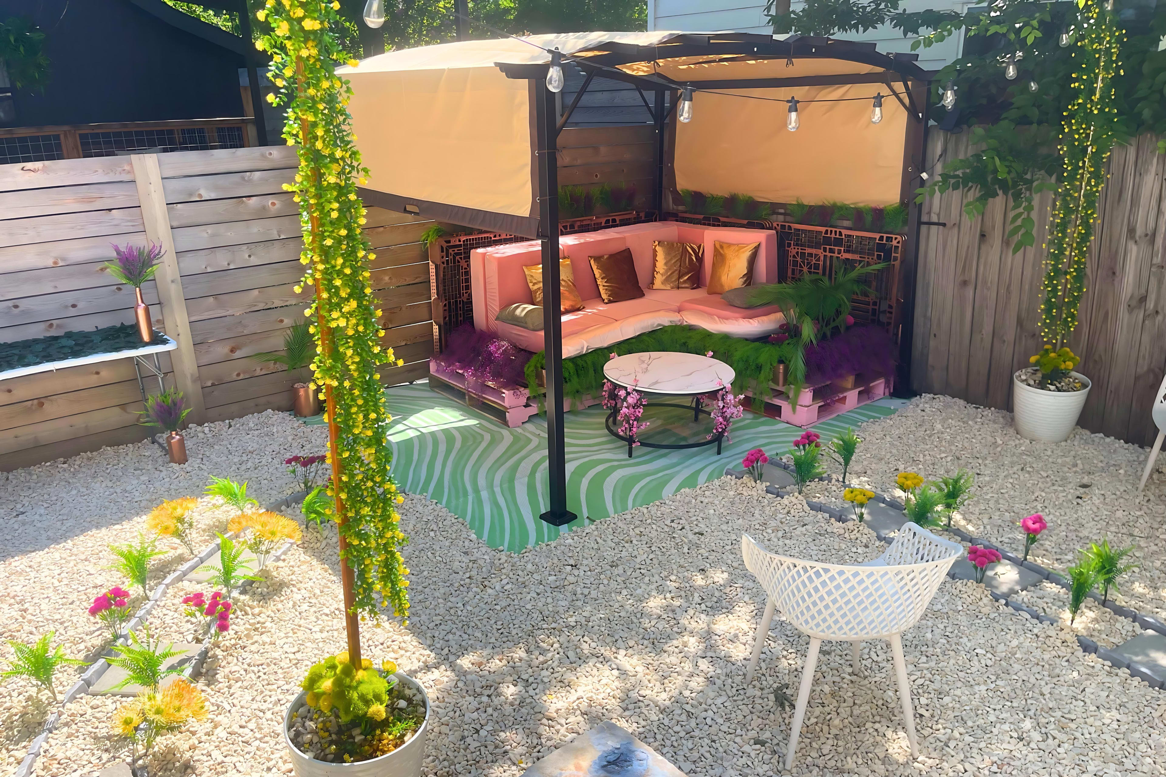 The image shows a landscaped outdoor space featuring a covered seating area with pink cushions, a round table, and decorative plants arranged on gravel.