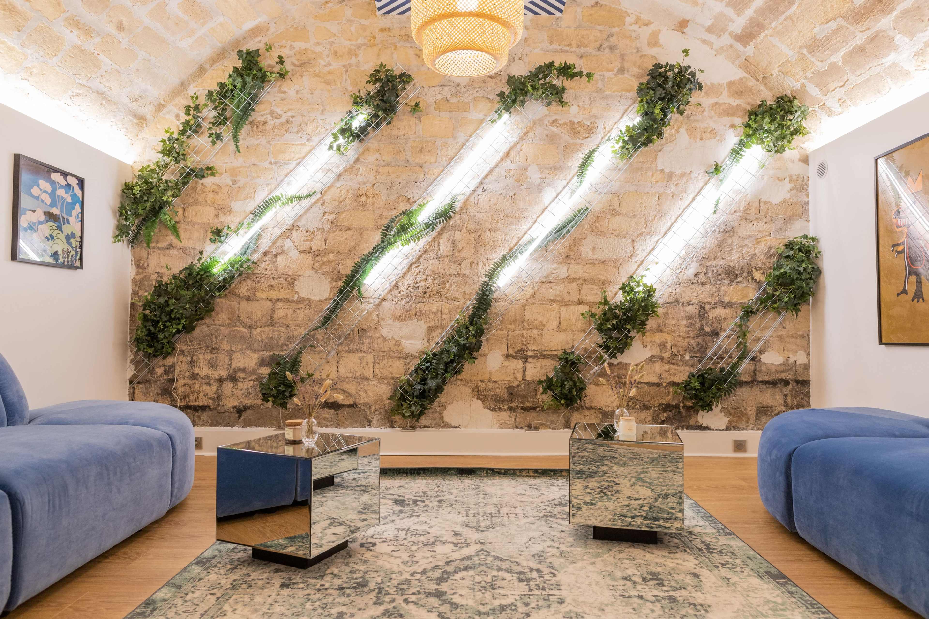 The image shows a modern lounge area featuring blue sofas, mirrored coffee tables, and a stone wall adorned with vertical greenery illuminated by LED lights.