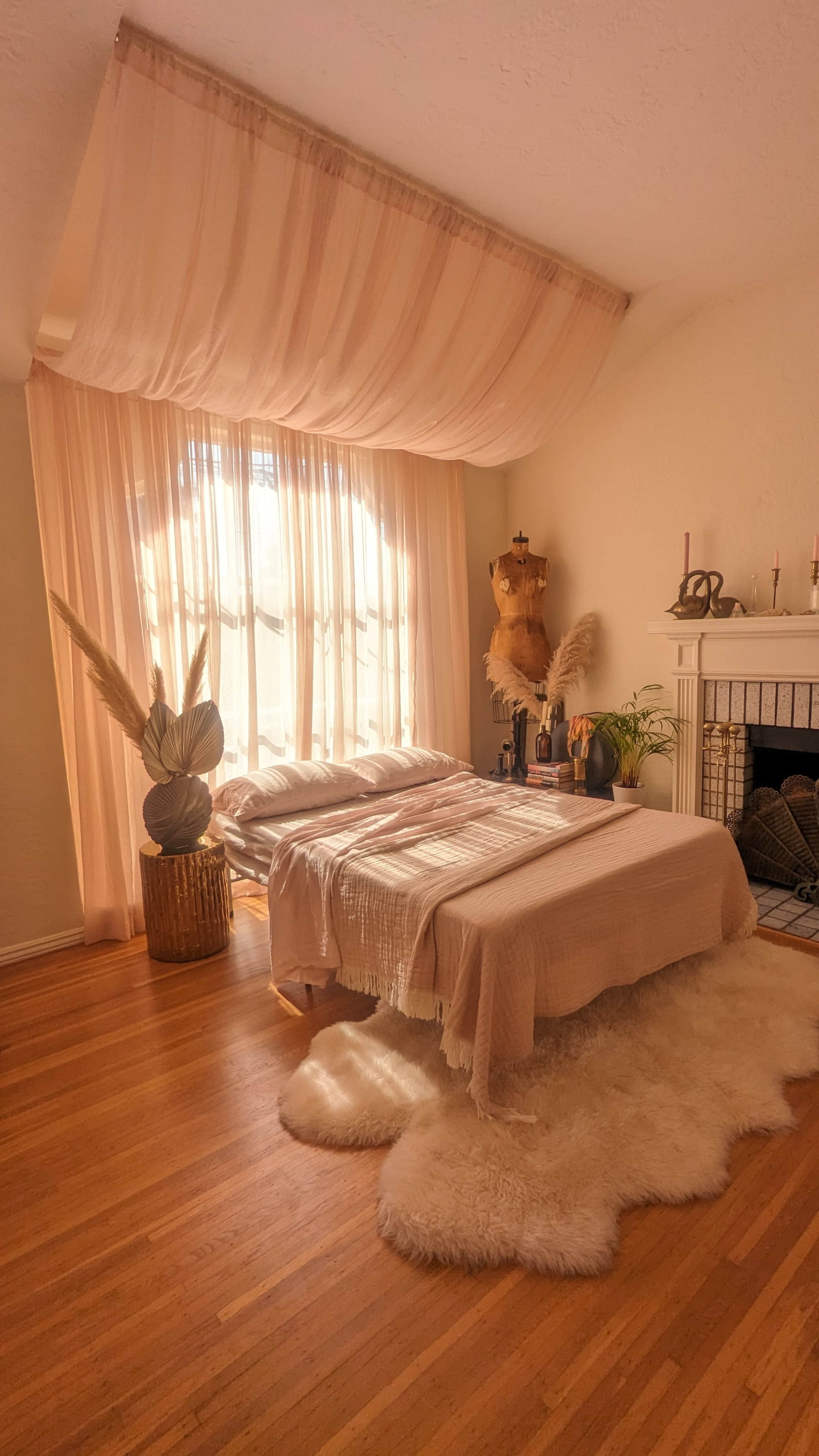 The room features a bed draped with a light-colored blanket, surrounded by plants, a fireplace, and large windows covered with sheer curtains.
