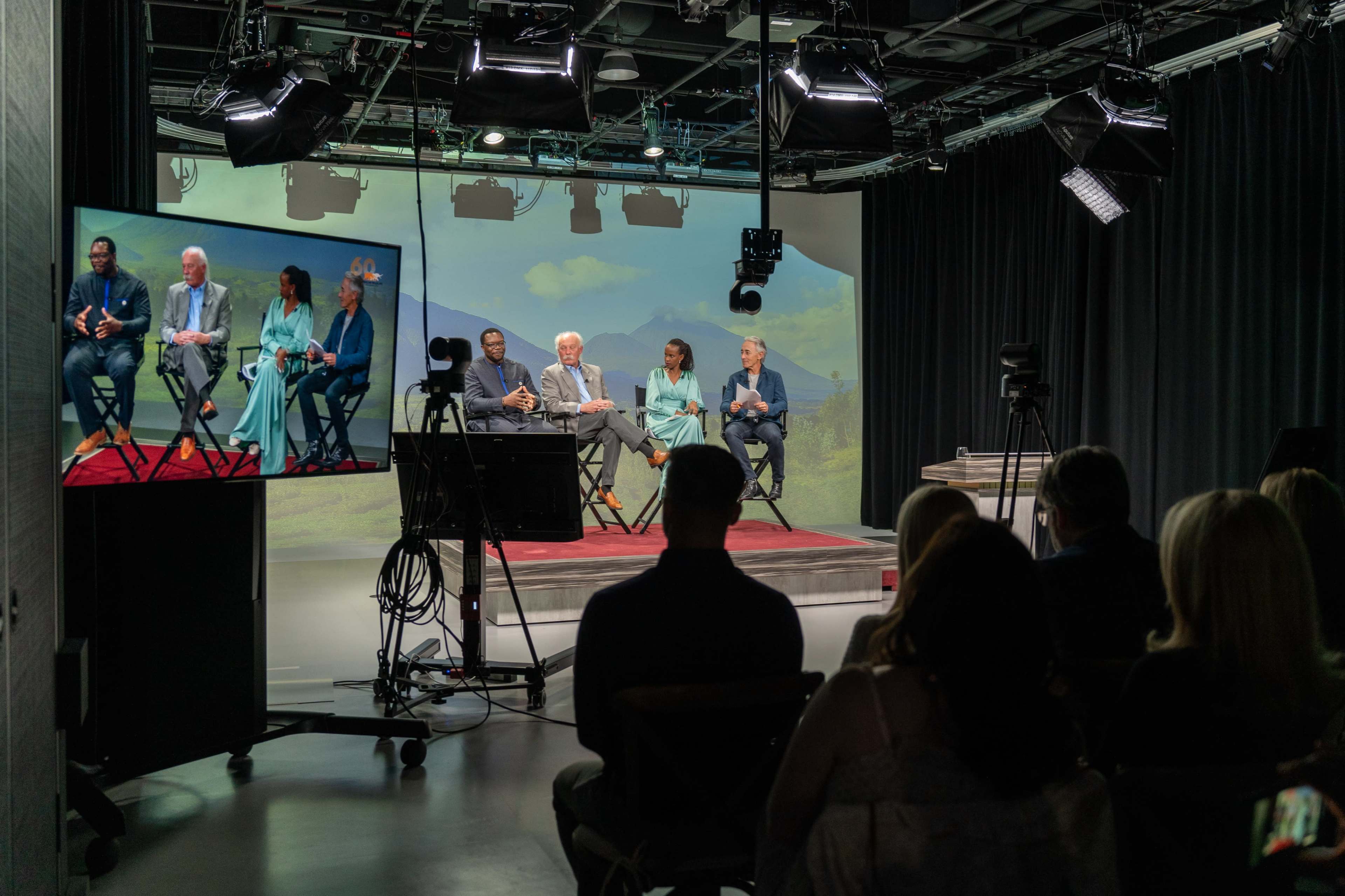A panel discussion is taking place on a stage with five speakers seated in front of a large screen displaying a scenic background, while an audience is visible in the foreground.