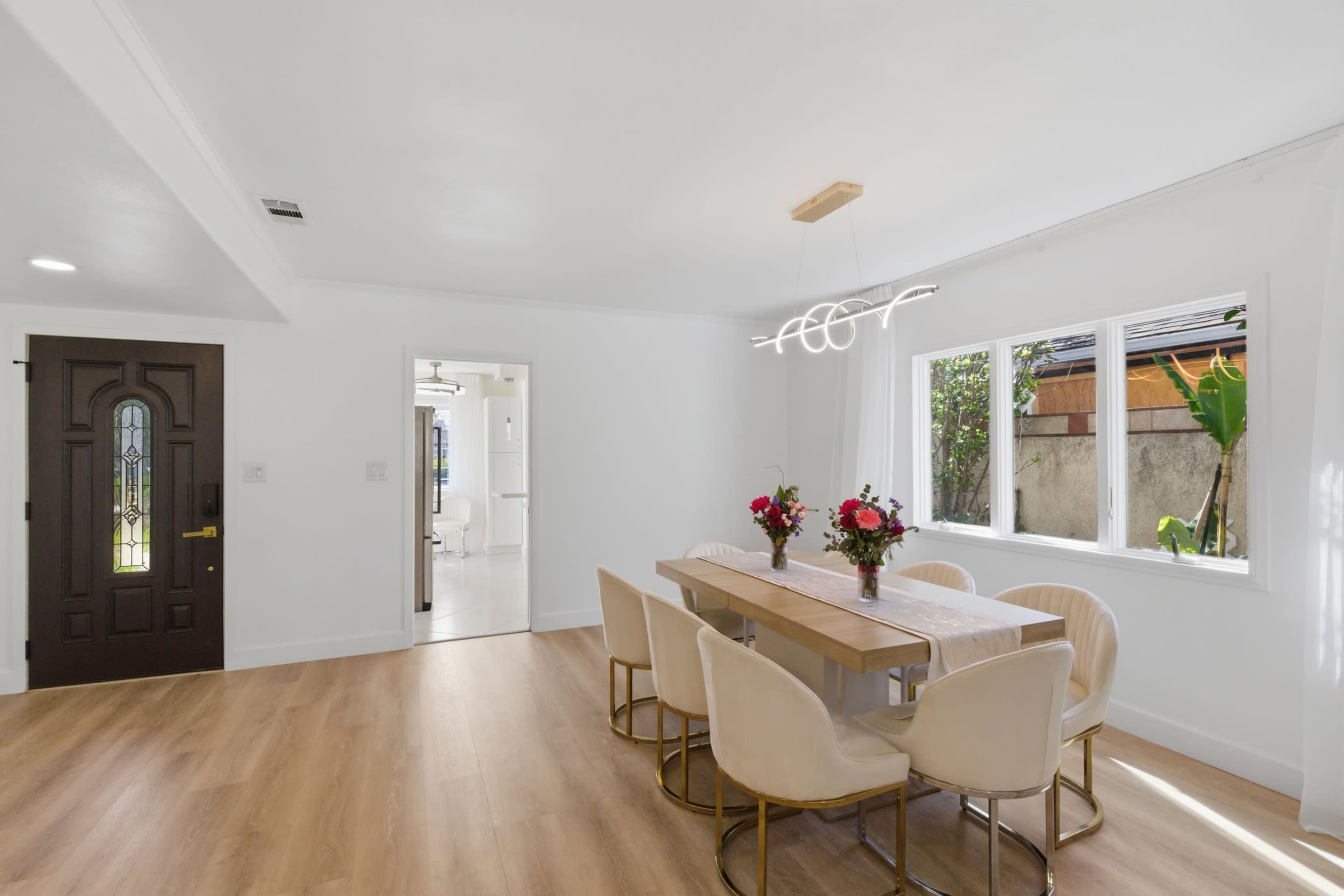 A modern dining area features a wooden table surrounded by beige chairs, adorned with floral centerpieces, and is illuminated by a contemporary light fixture.