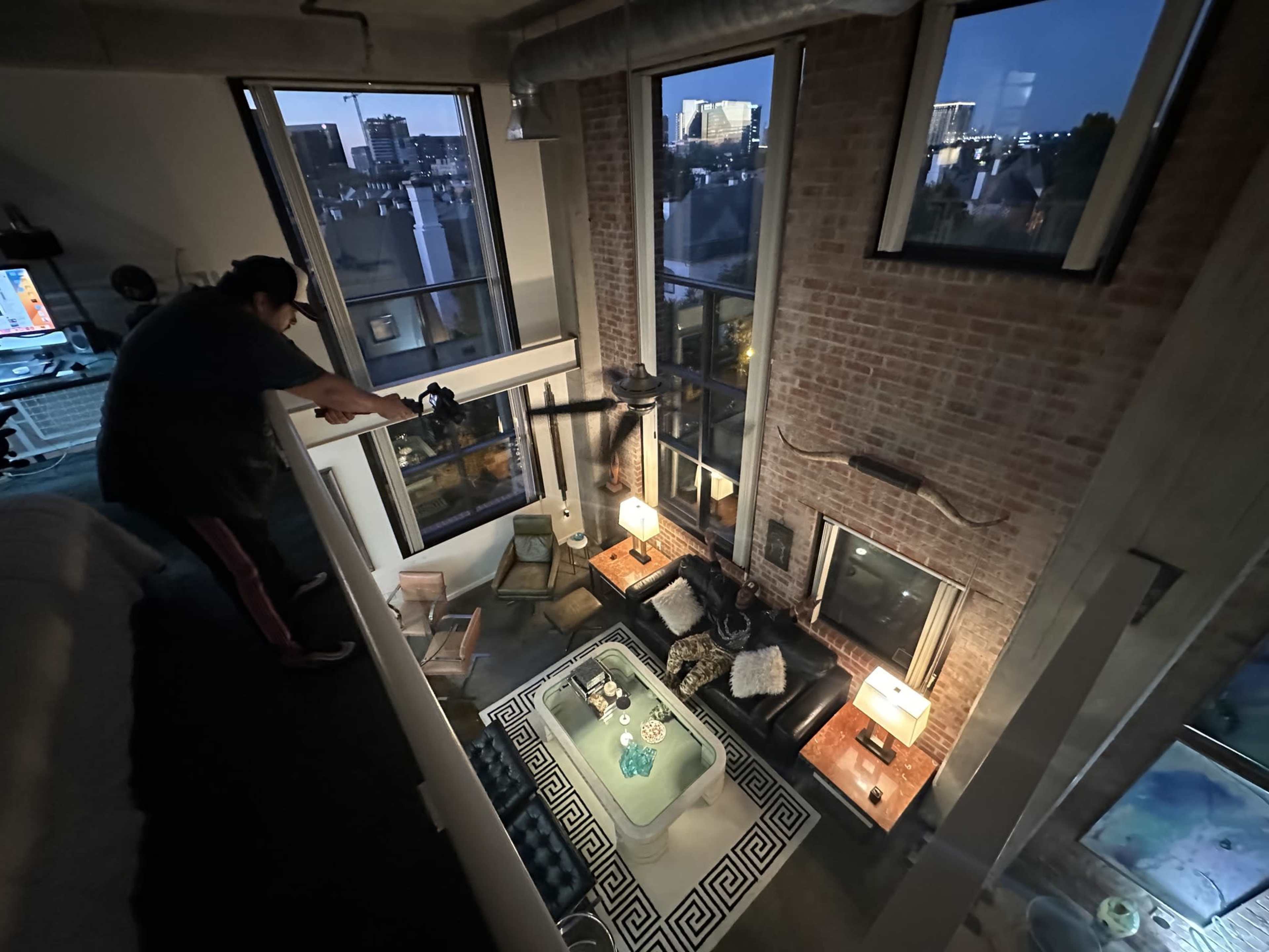 A person leans over a railing in a loft apartment, overlooking a living area with large windows, two lamps, and a coffee table.