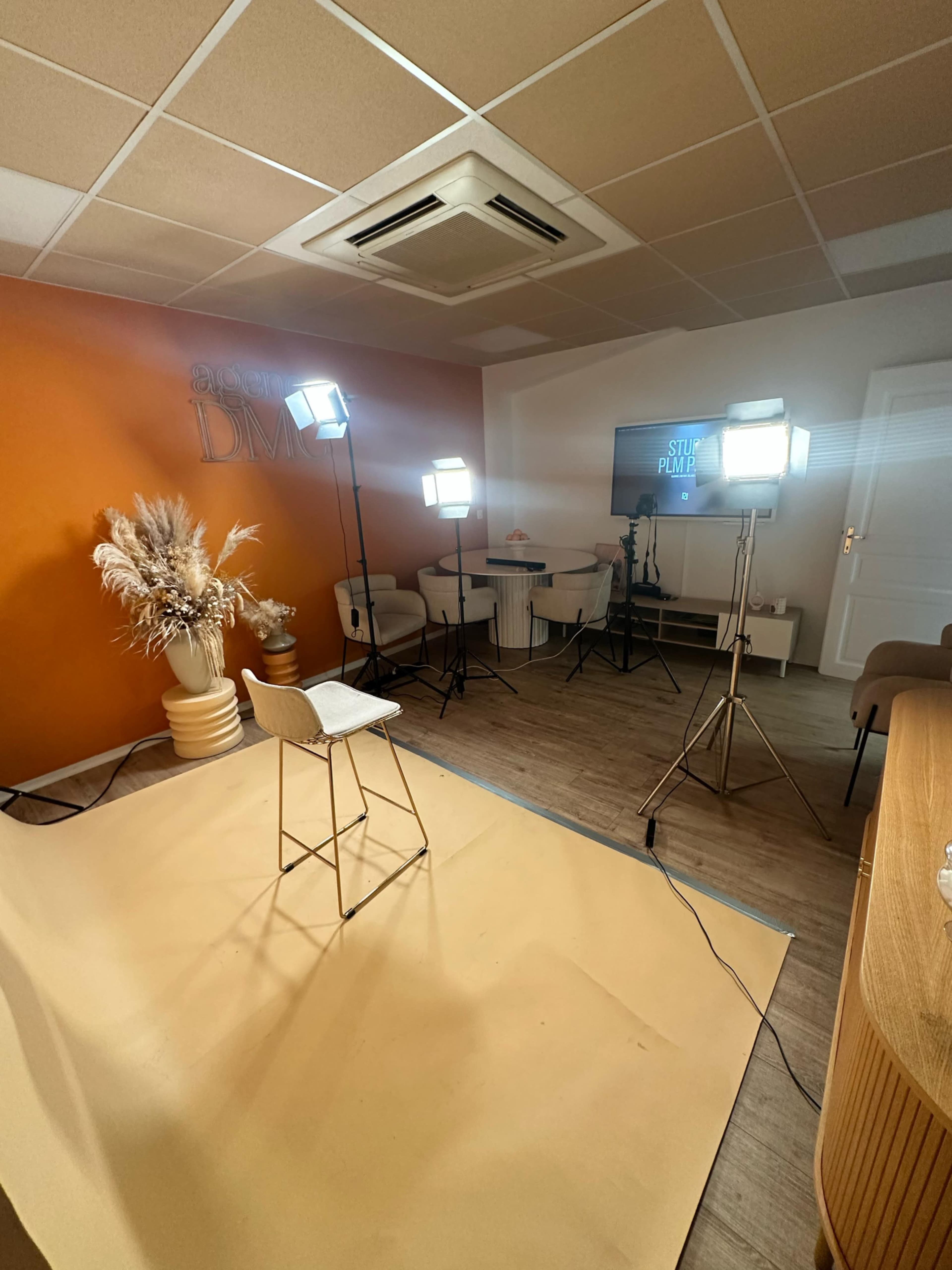 The image shows a well-lit studio setup with several light stands, a stool on a beige floor, and a backdrop featuring the words “SALEM DM” on an orange wall.