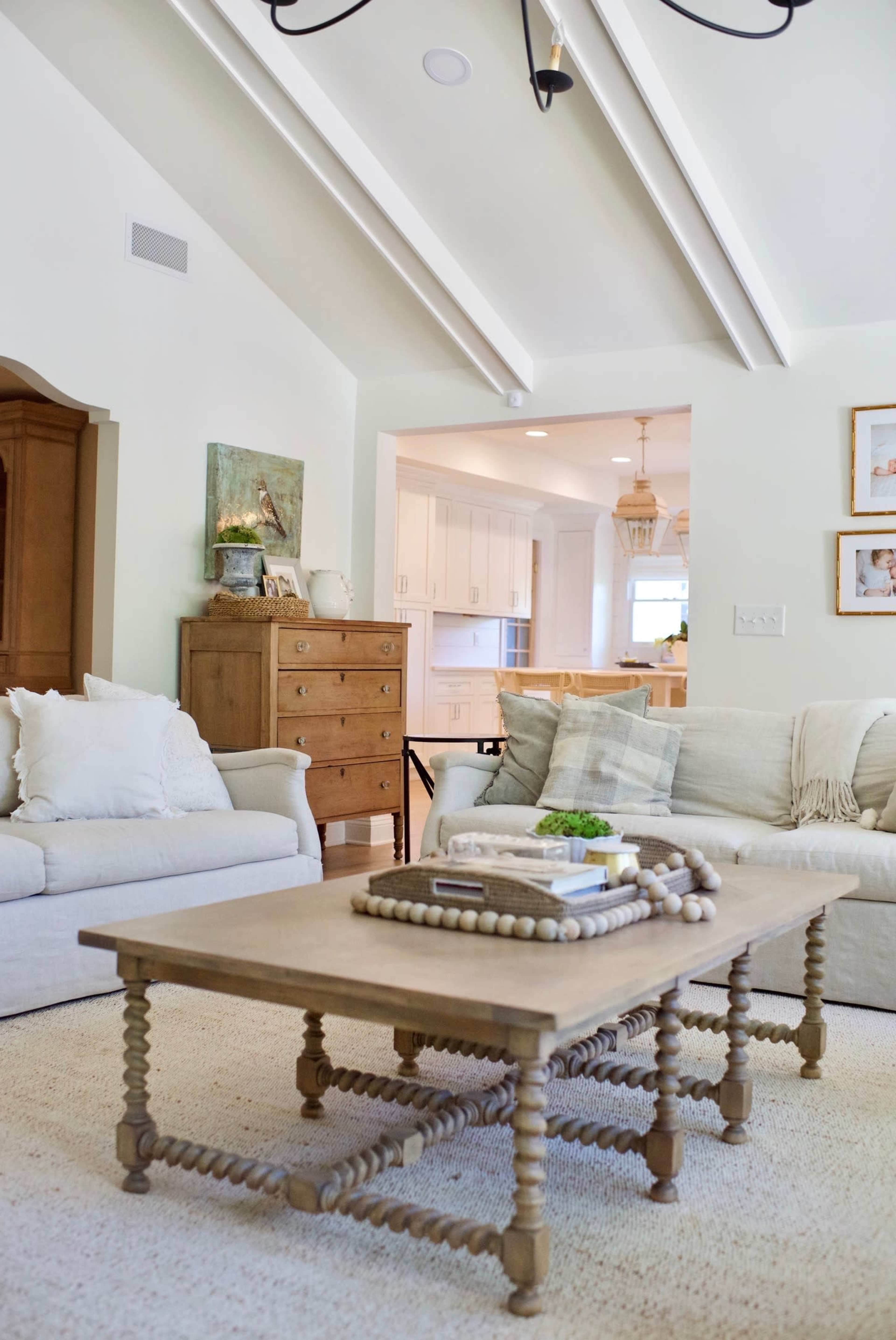 A light-filled living room features a beige sofa, a wooden coffee table with decorative items, and a view into a kitchen area.