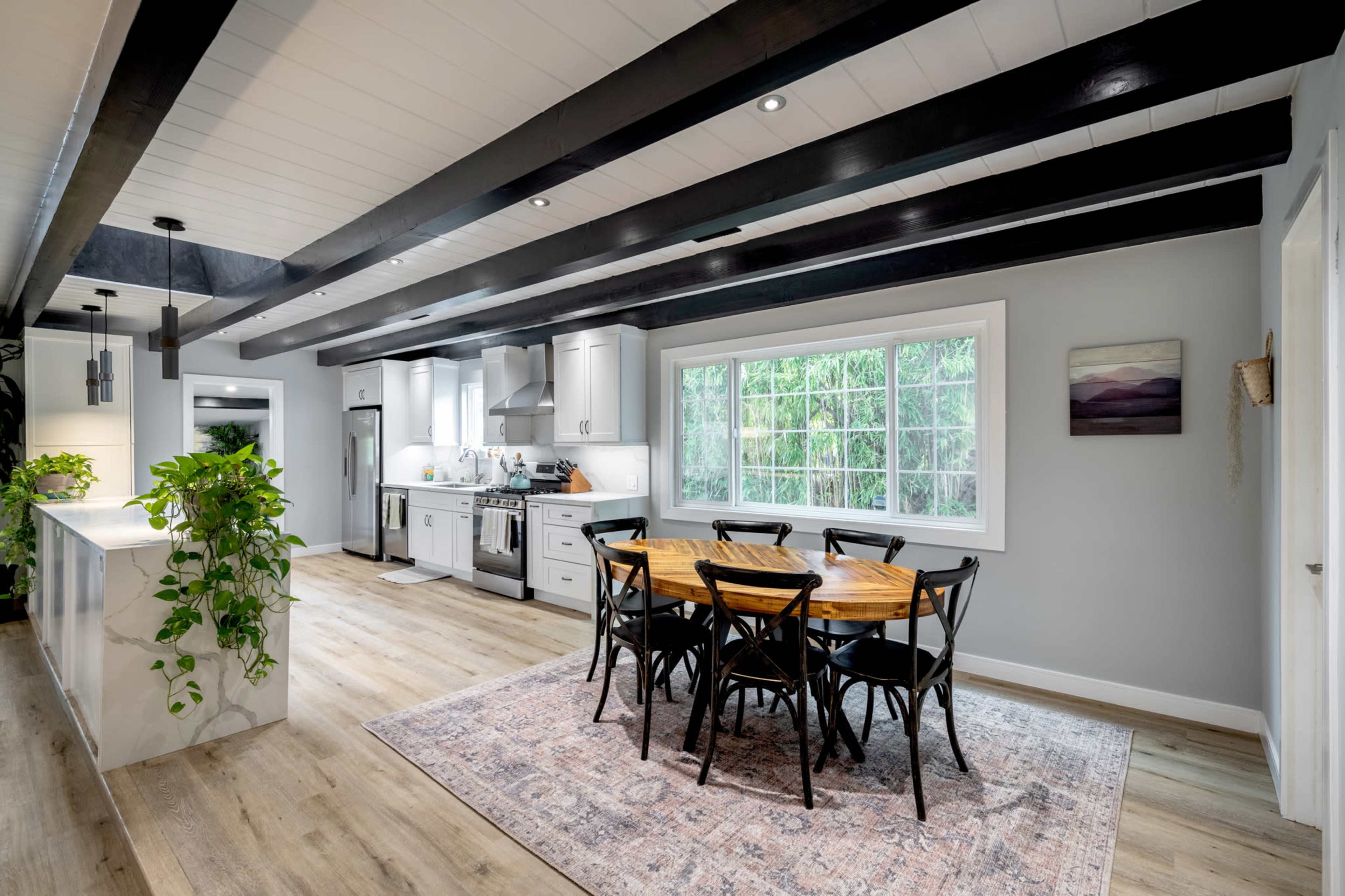 The image shows a modern kitchen and dining area featuring a wooden dining table surrounded by black chairs, with a large window allowing natural light and views of greenery outside.