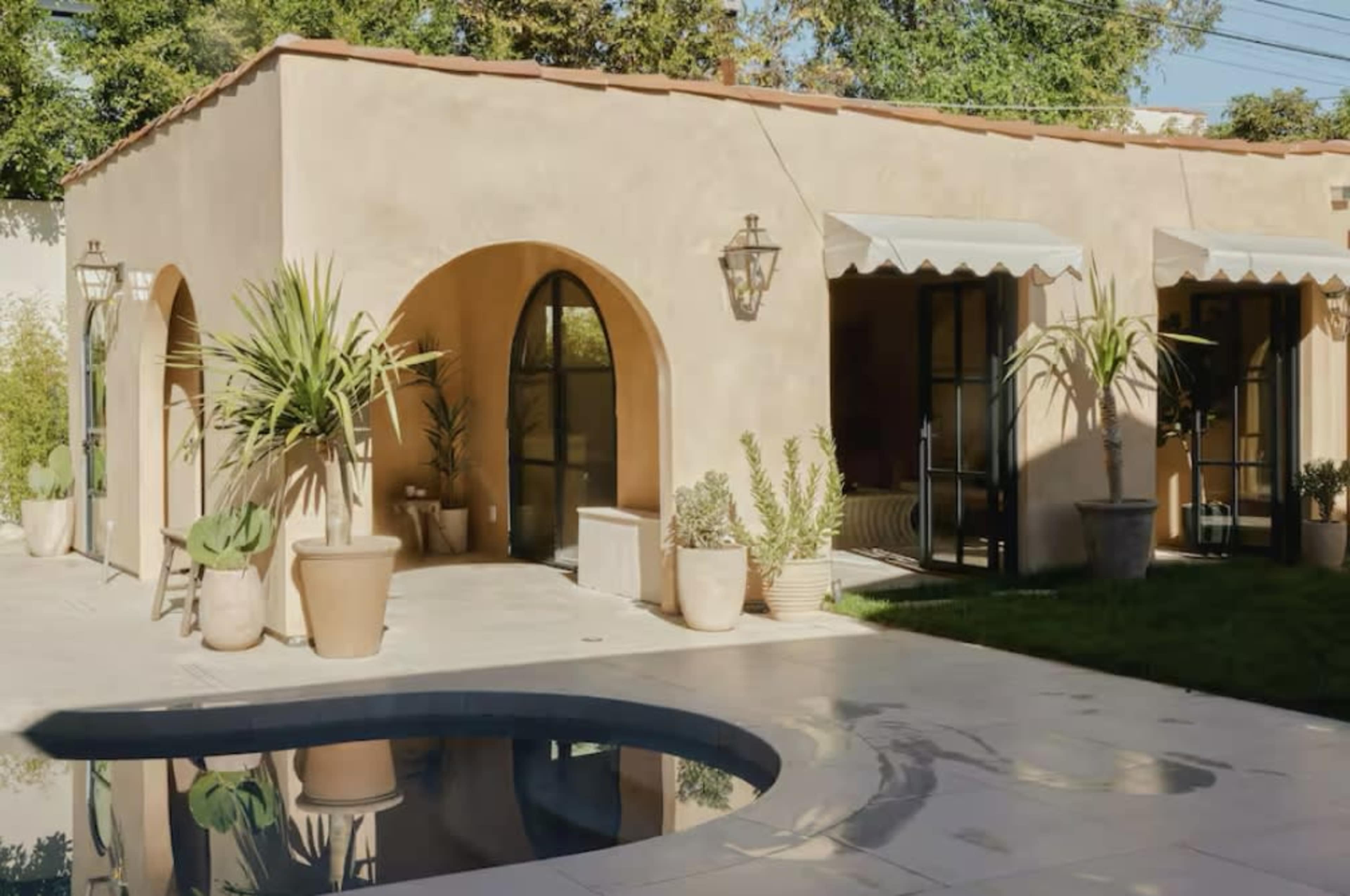 The image shows a small, stucco building with arched windows and doorways, surrounded by potted plants and a swimming pool in the foreground.