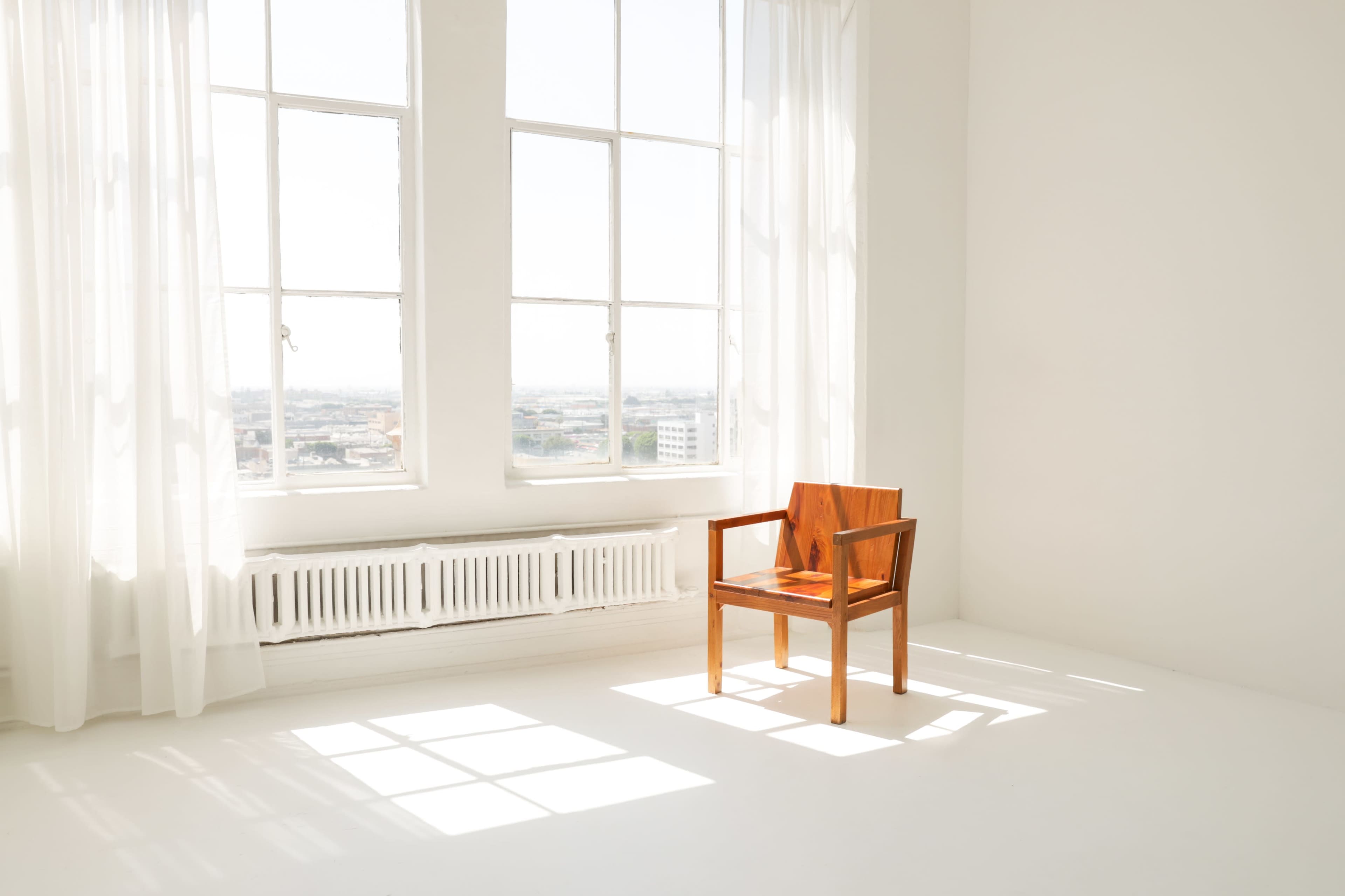 A wooden chair is placed in a bright, empty room with large windows that let in sunlight.