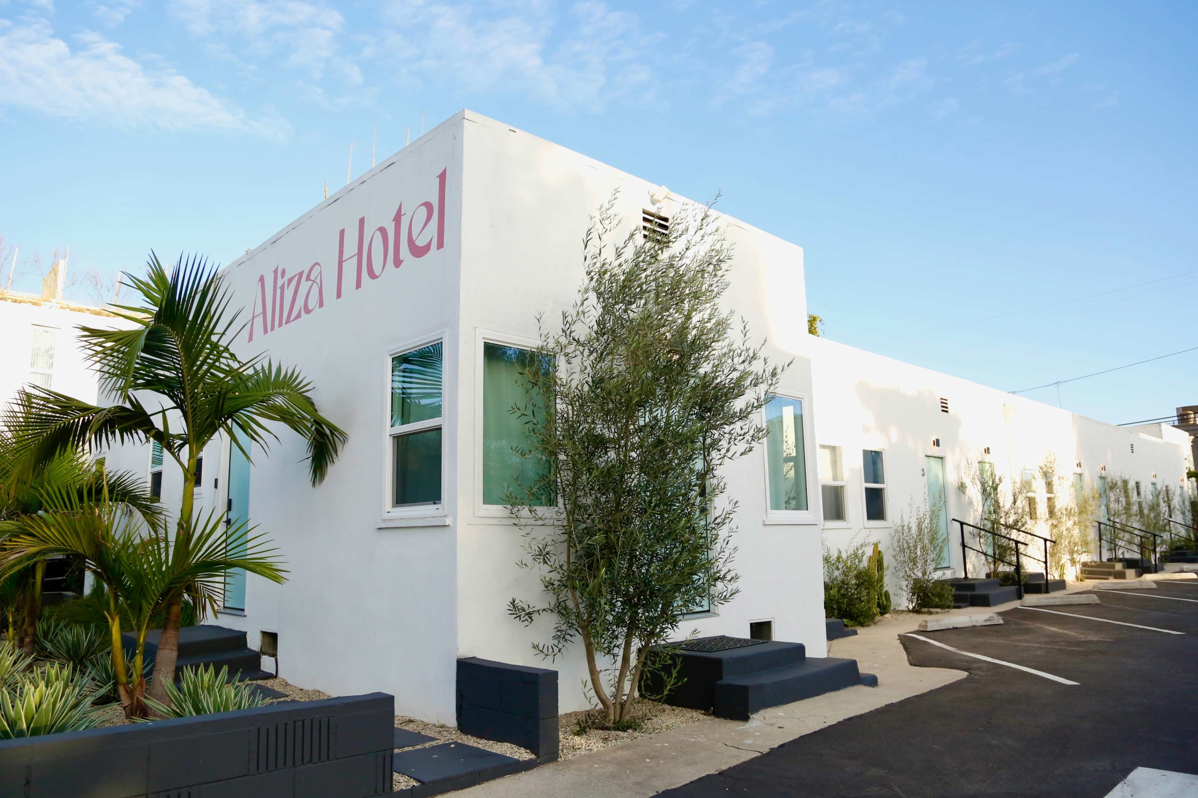 The Aliza Hotel features a modern, white building with landscaping and parking spaces in a sunny outdoor setting.