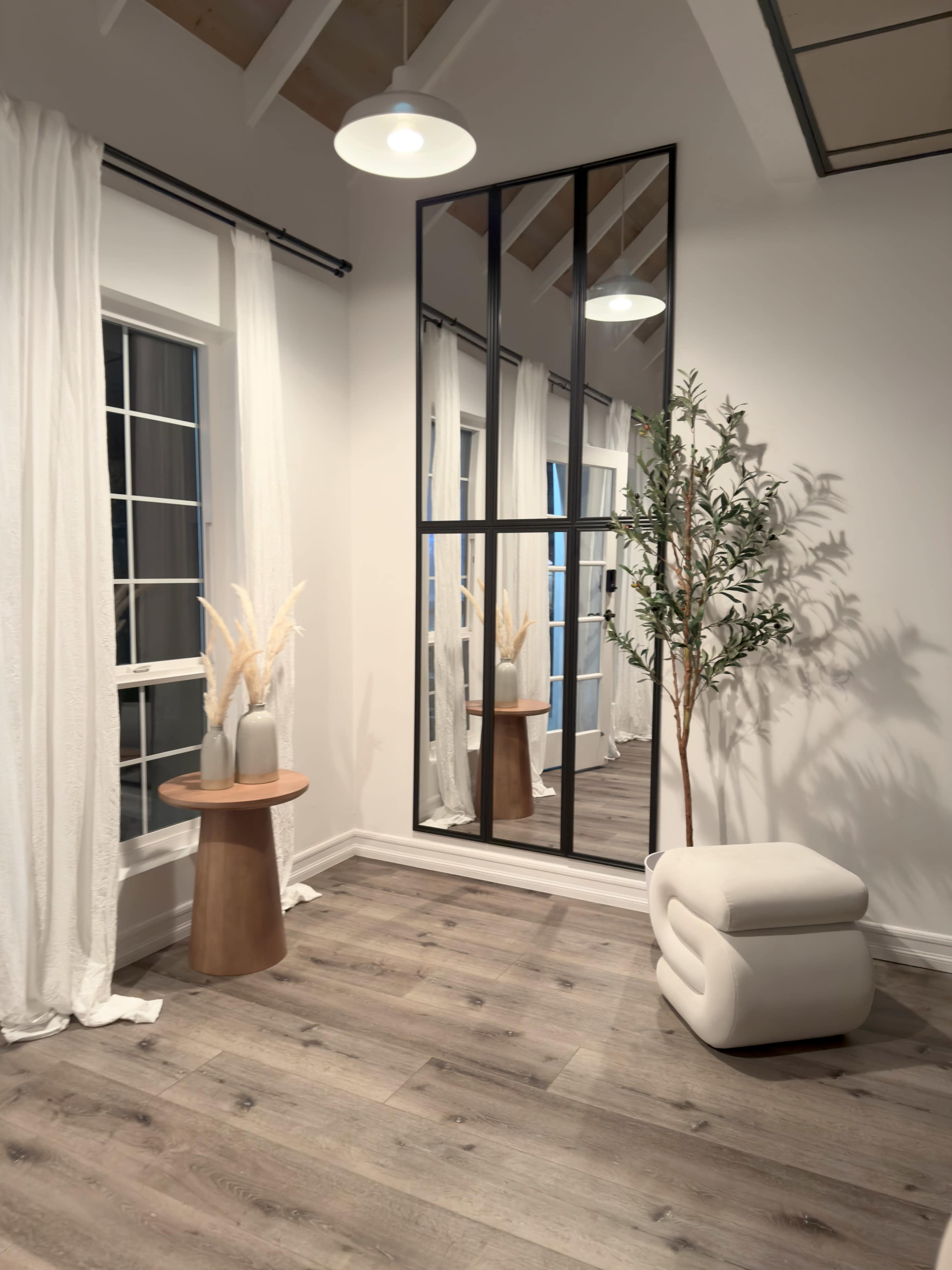 The image shows a bright interior space featuring a large mirror, a small wooden table with decor, a potted plant, and white curtains framing a window.