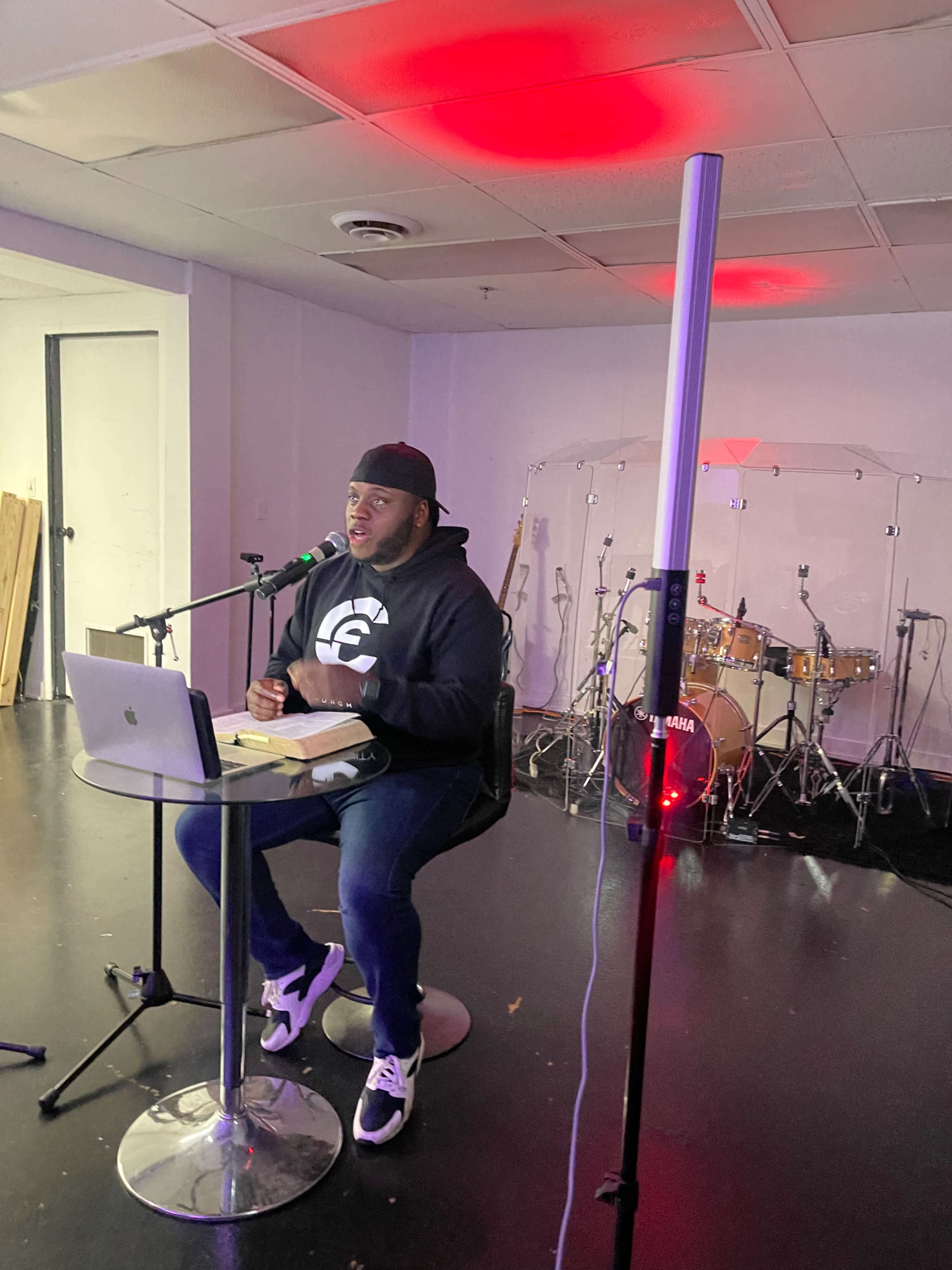 A person sitting at a small table with a laptop and microphone in a room with musical instruments and colored lights.