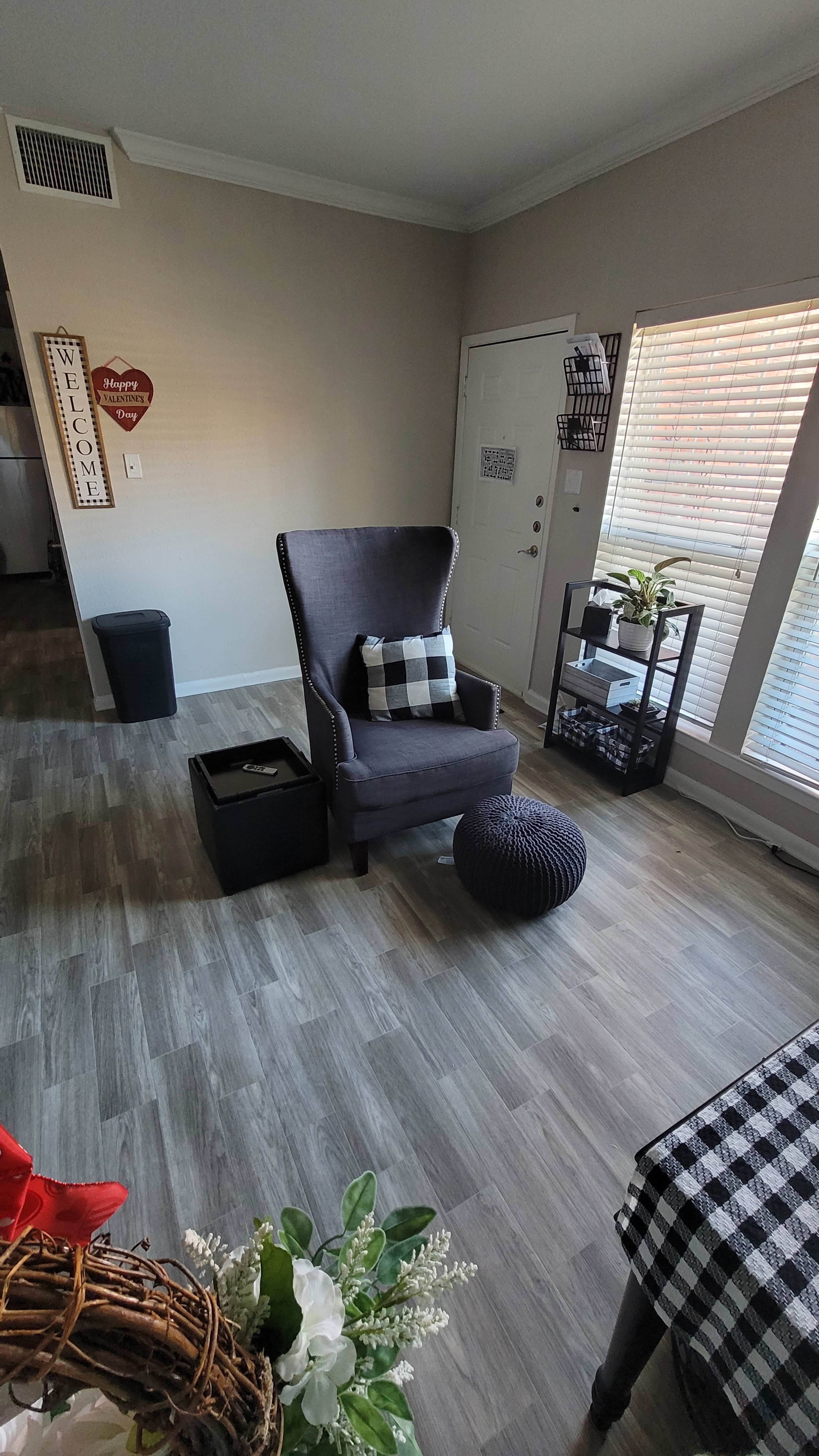 The scene shows a cozy living room with a gray wingback chair, a small pouf, a decorative plant near a window, and a door leading outside.