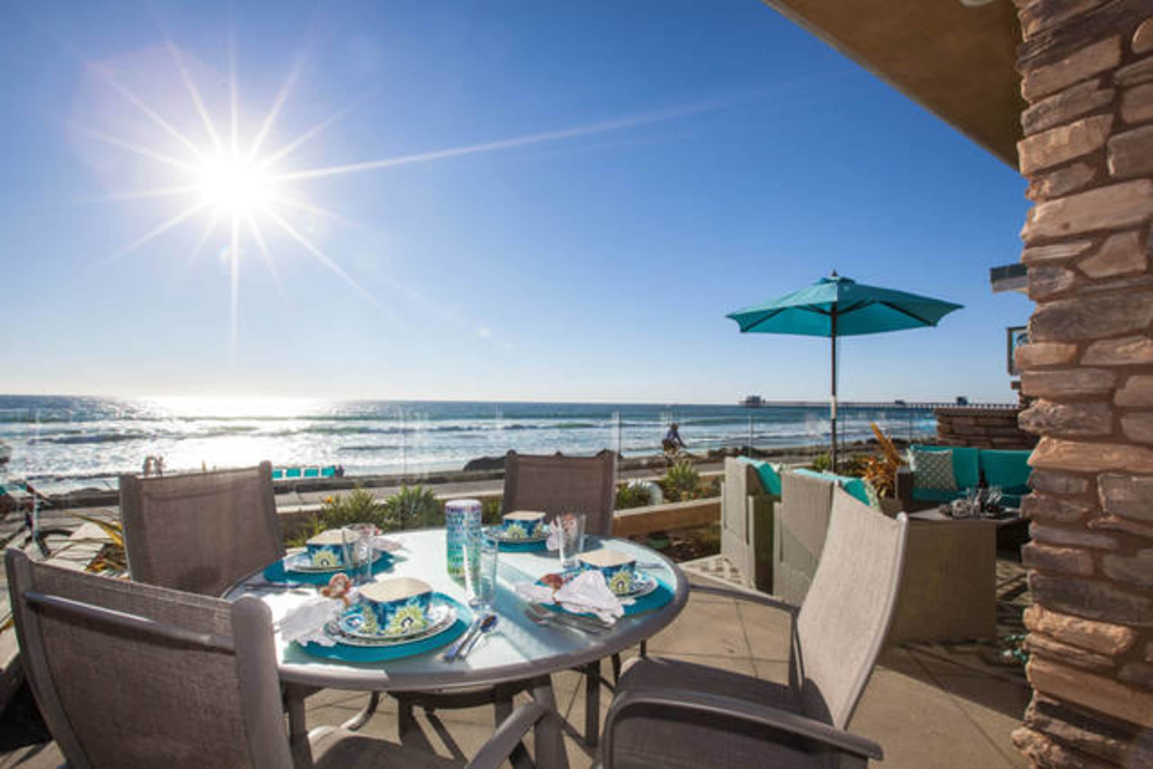 A patio dining set is positioned near the beach, overlooking the ocean with sun shining brightly in the background.