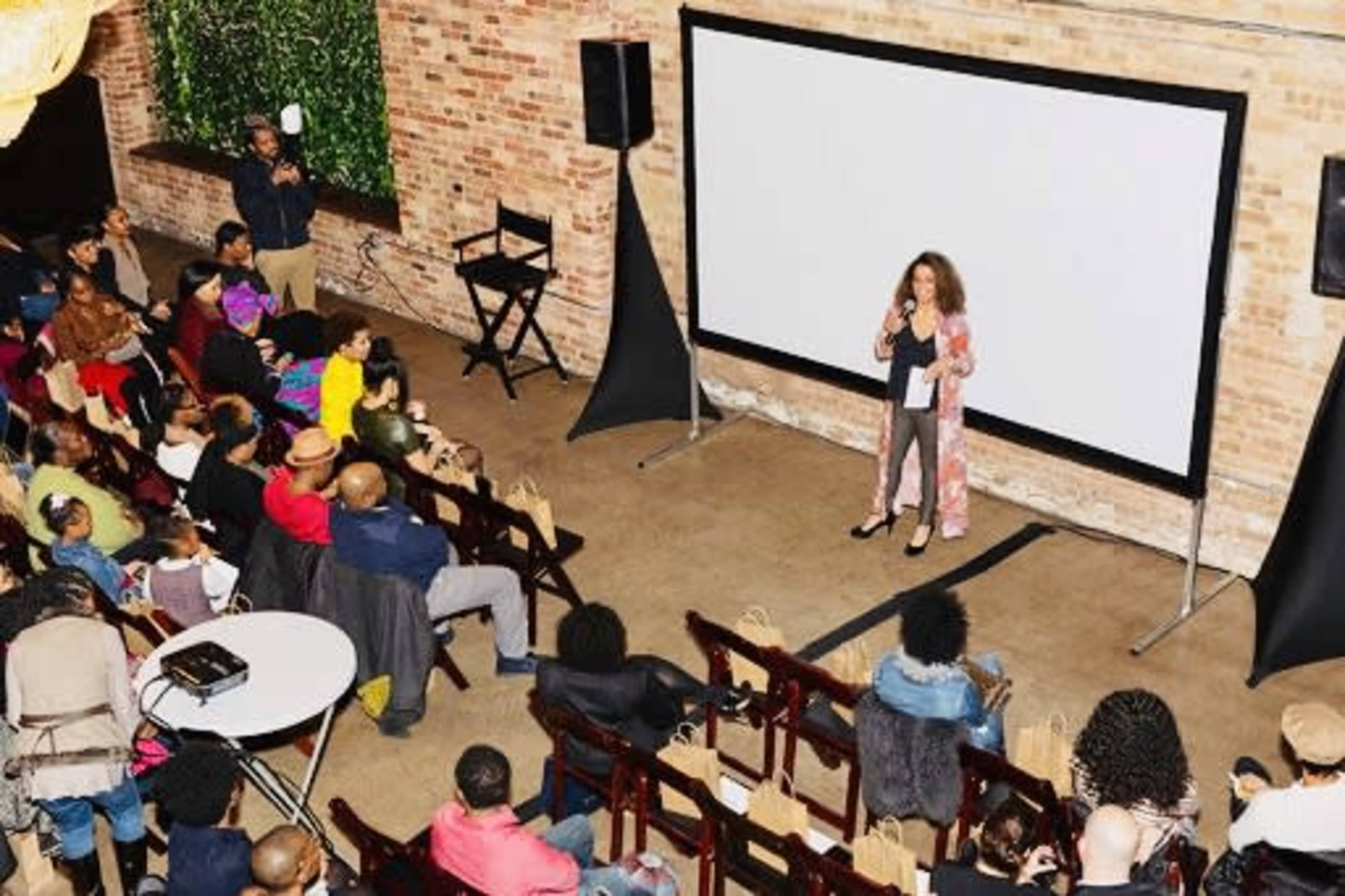 A speaker stands on stage in front of a large screen while an audience sits in rows of chairs facing her during an event in a brick-walled venue.