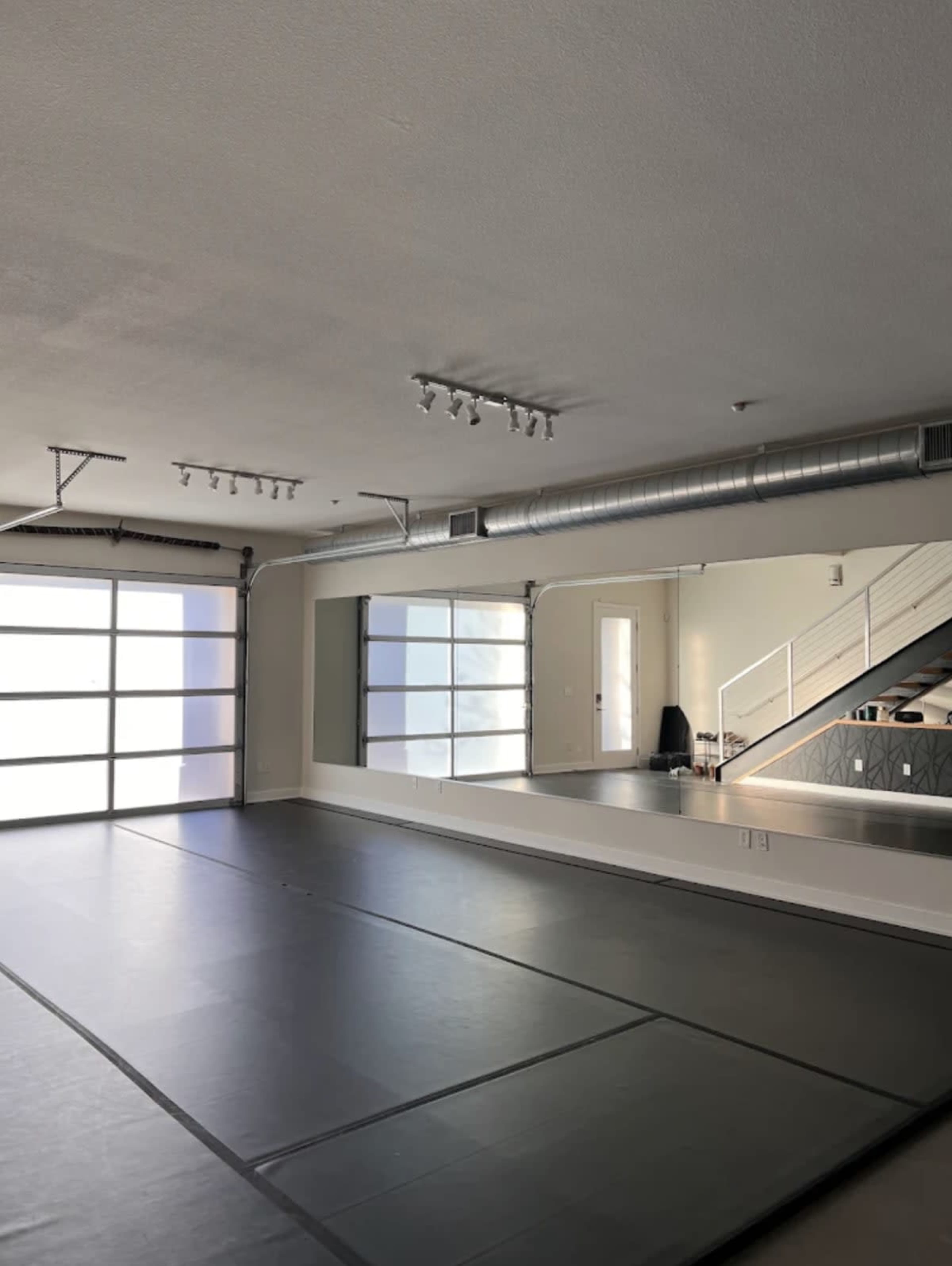 The image shows a spacious, empty room with a mat-covered floor, a large mirror on one wall, and industrial-style lighting overhead.