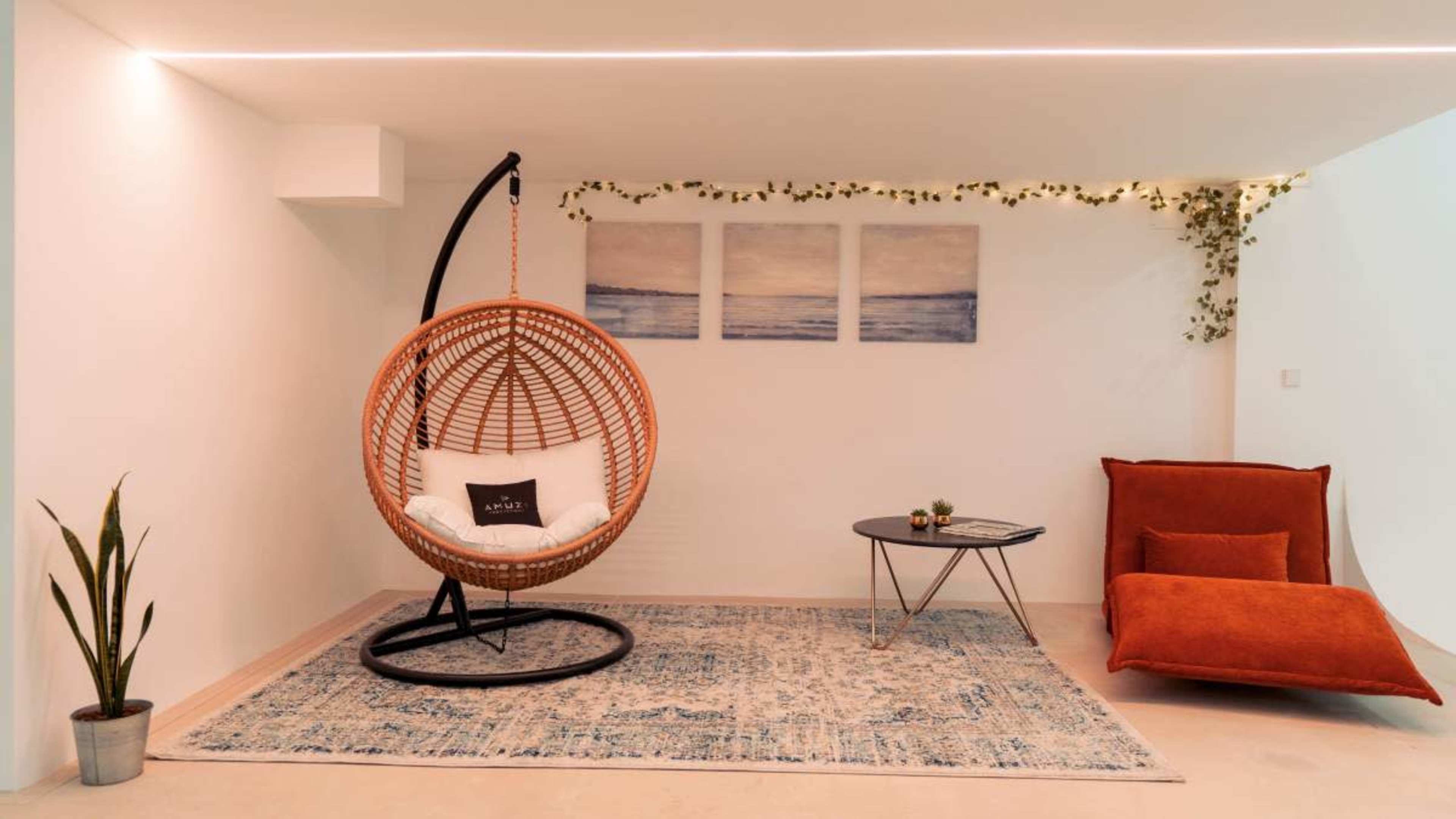 The image shows a modern interior space featuring a hanging rattan chair, a sleek coffee table, and a vibrant orange lounge chair on a decorative rug.