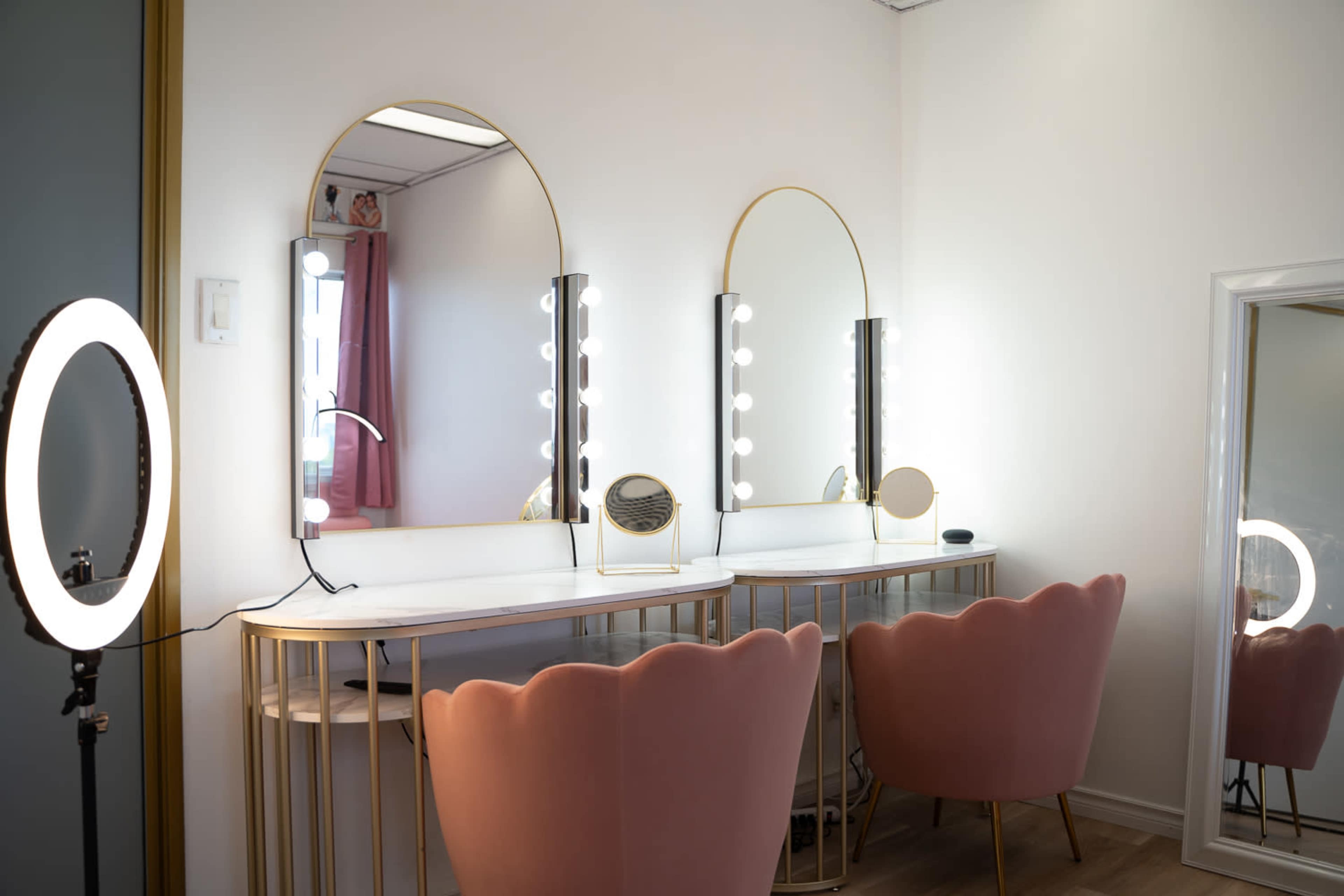 The image shows a modern vanity area with two pink upholstered chairs and a white table featuring large mirrors surrounded by light bulbs.