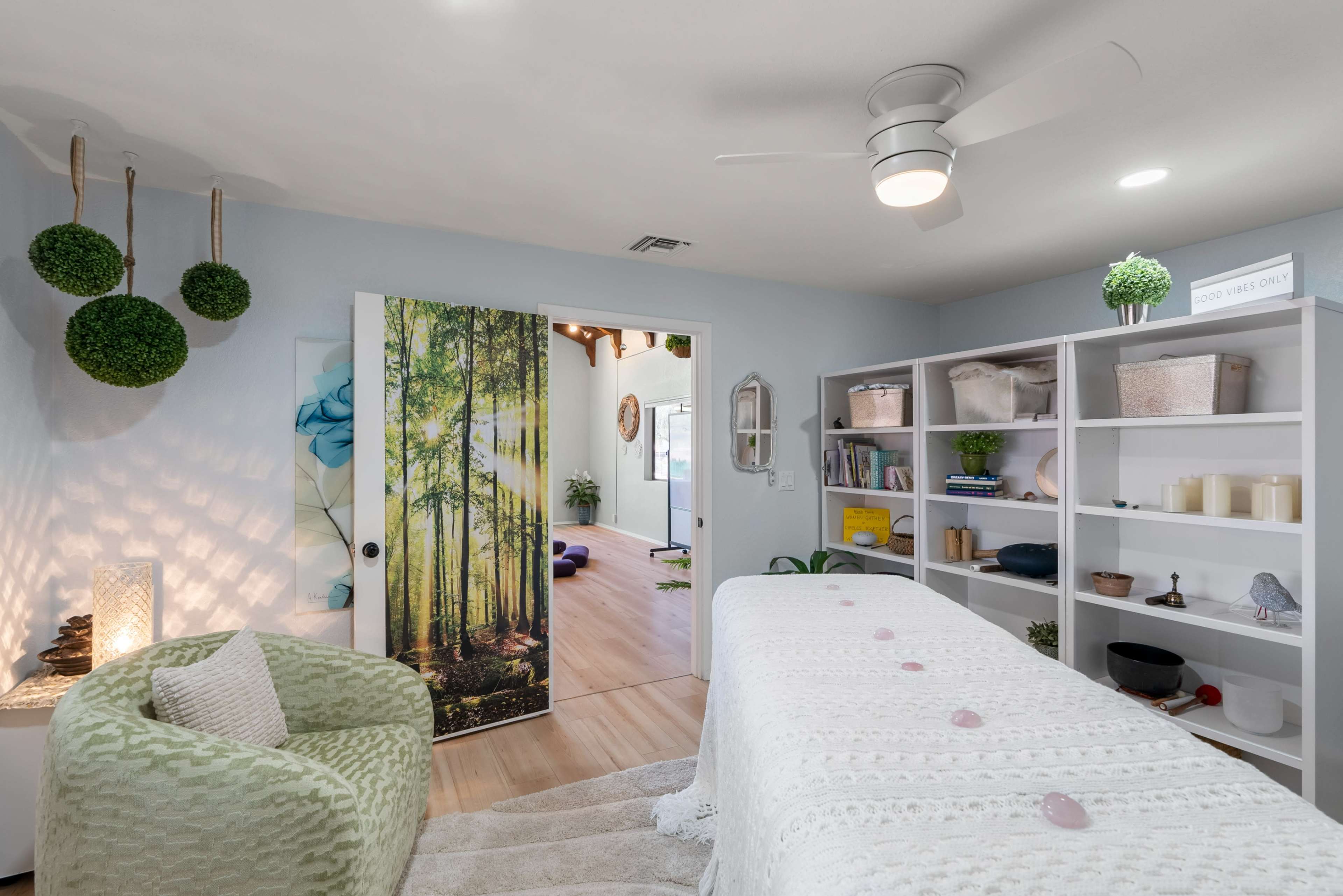 A serene therapy room features a massage table adorned with stones, surrounded by shelves of organized wellness items and a mural of a forest on the door.