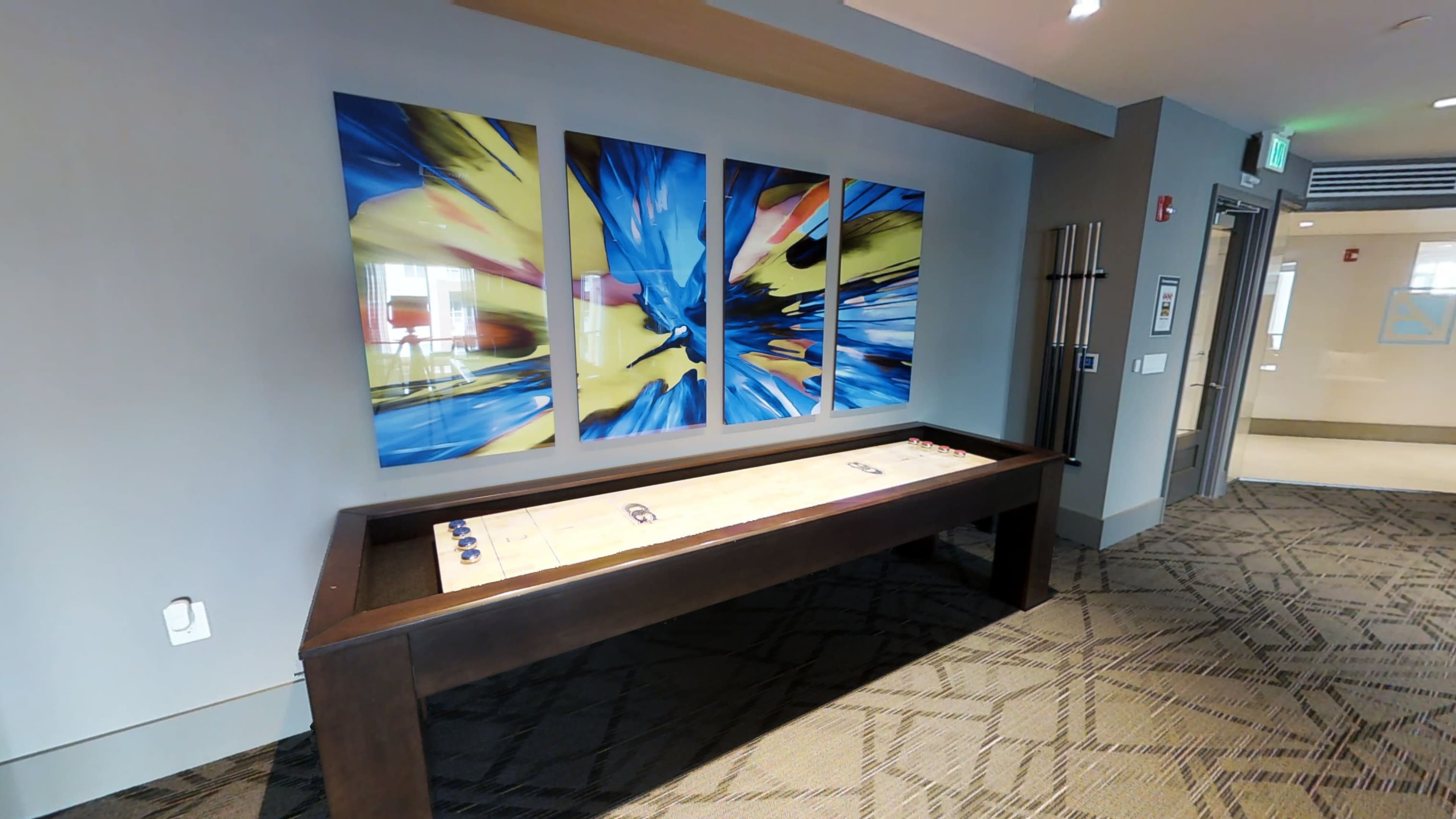 A modern room with a shuffleboard table against a wall adorned with three colorful abstract art pieces.