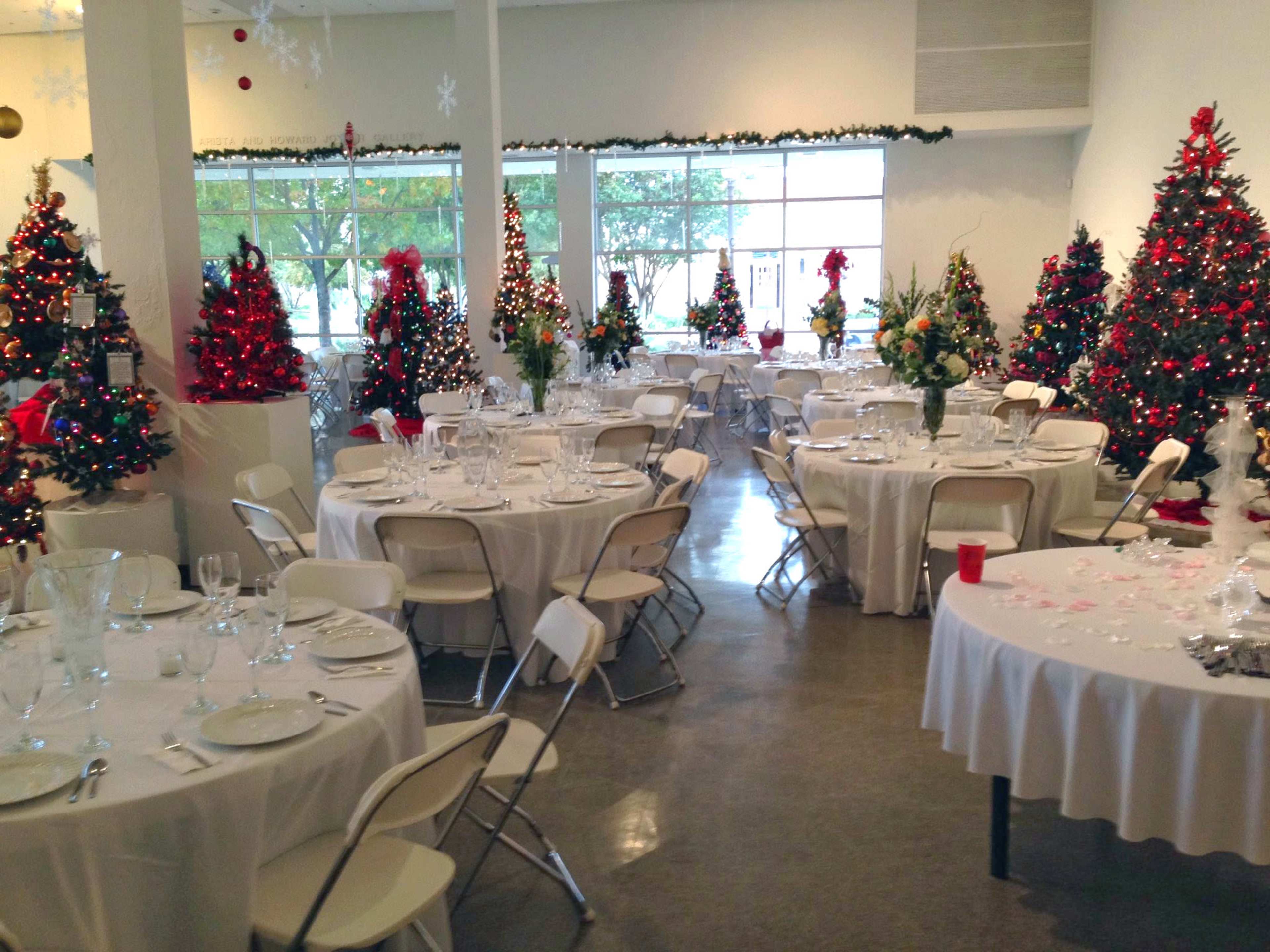 A decorated banquet hall with several round tables set for a meal, surrounded by Christmas trees adorned with lights and ornaments.