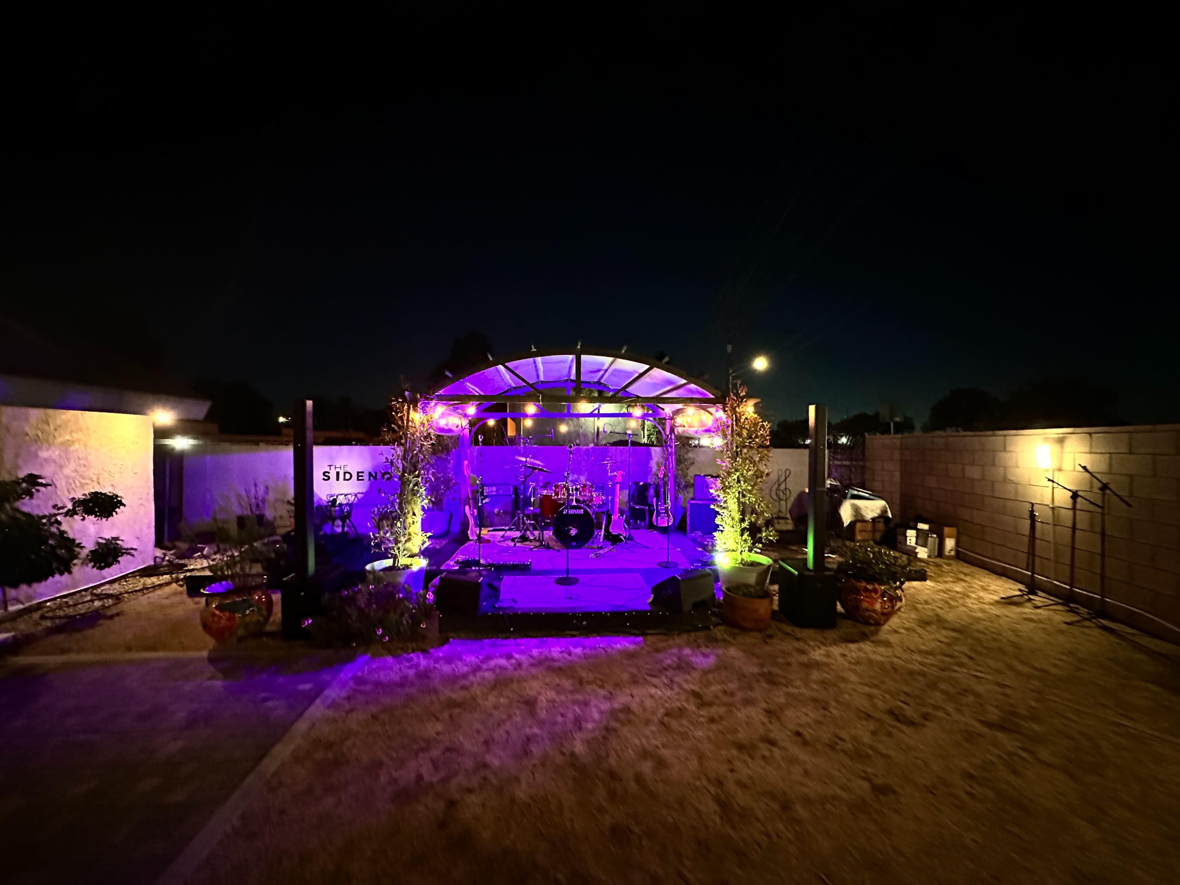 A stage set for a performance is illuminated by colorful lights in a dark outdoor space, surrounded by plants and a low wall.