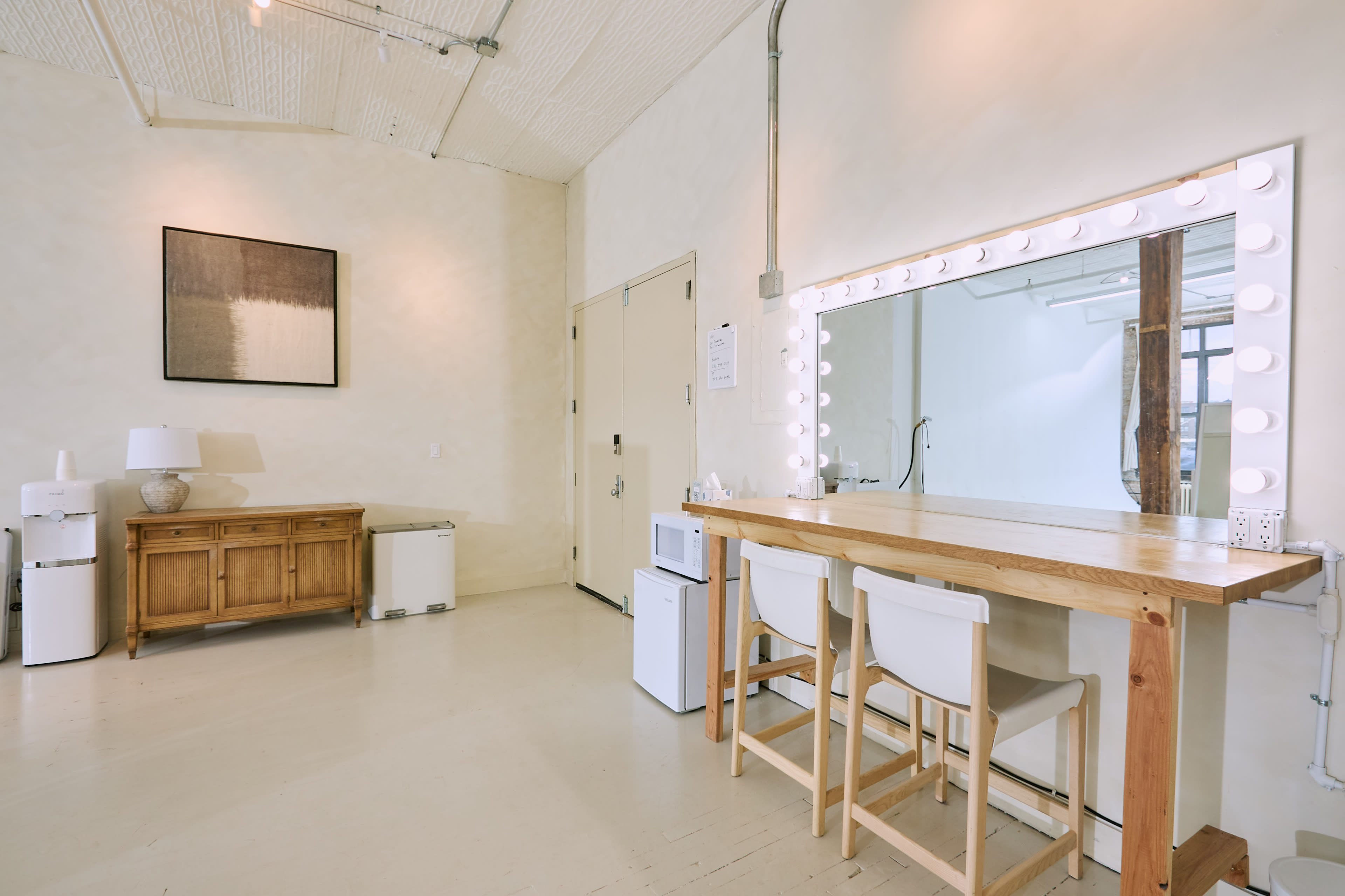 The image shows a minimalistic room featuring a wooden table with two white chairs, a mirror bordered by light bulbs, and a small kitchenette area with a lamp and appliances.