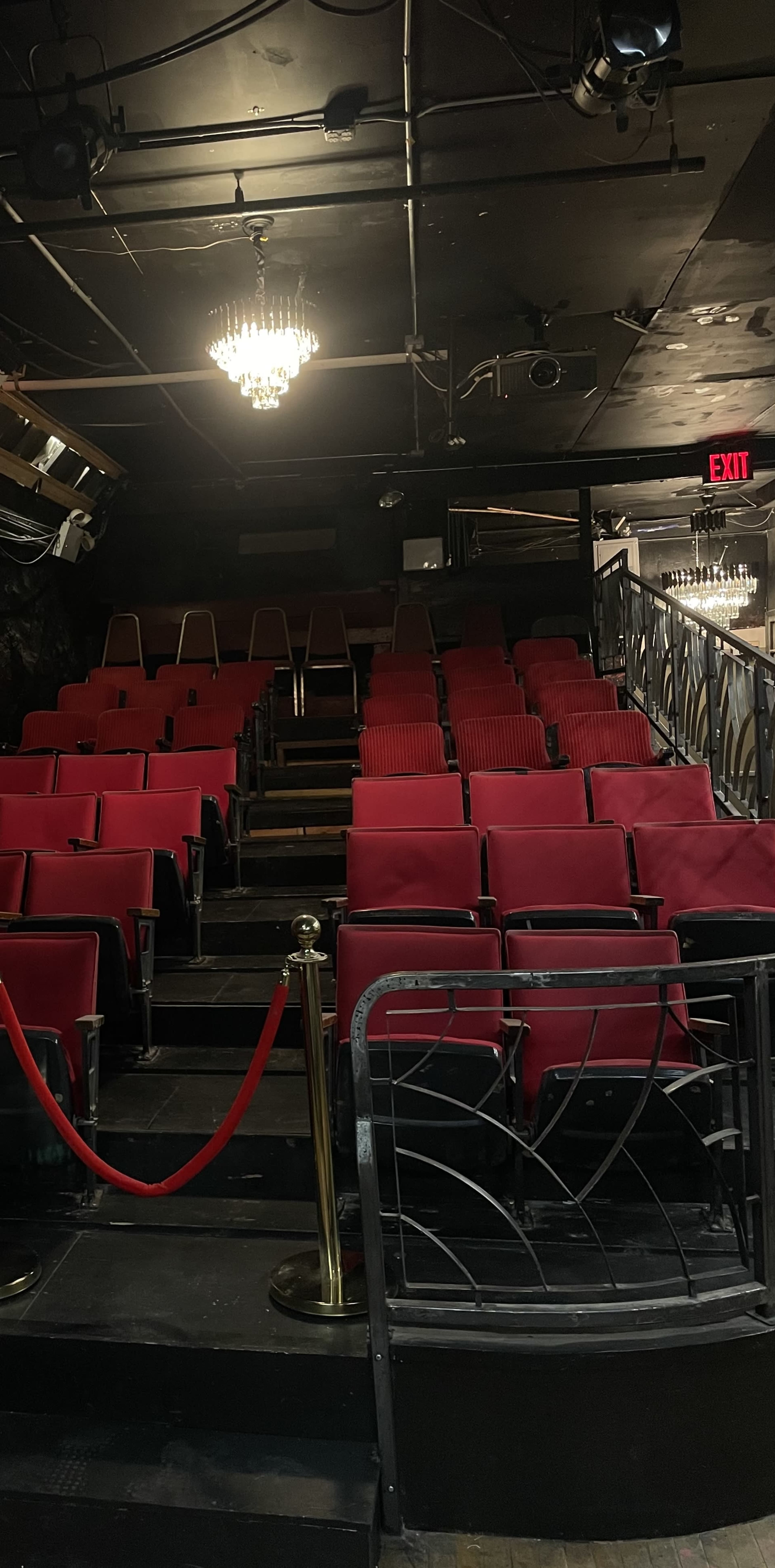 The image shows empty red theater seats arranged in a sloped seating area under dim lighting, with an exit sign visible in the background.