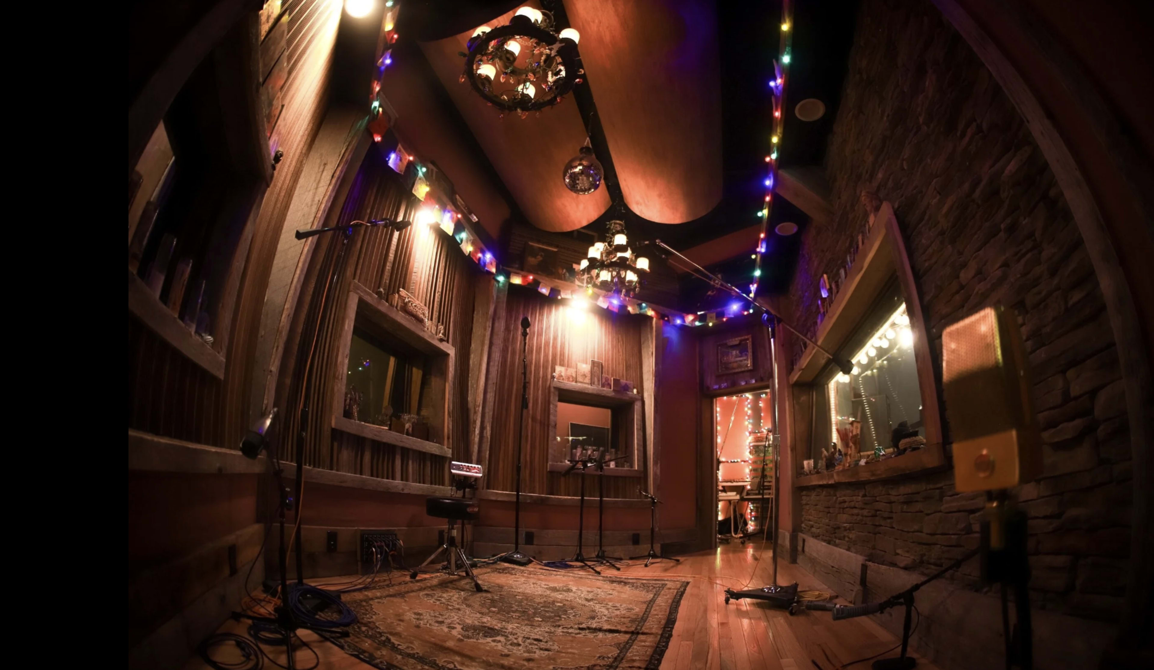 The image shows a cozy recording studio adorned with colorful string lights and wooden walls, featuring microphones and a large rug on the floor.