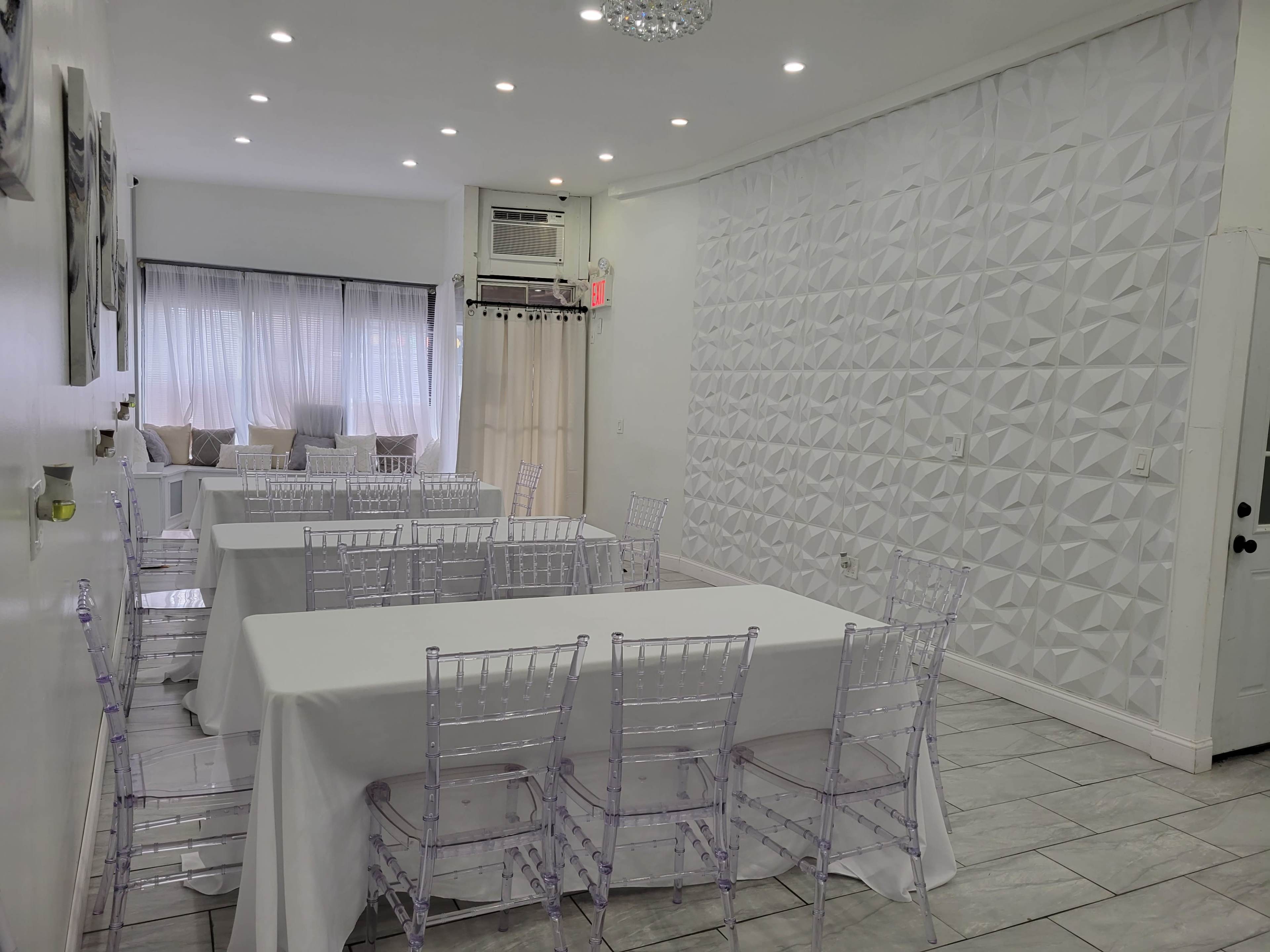 A spacious dining area with white tables covered in tablecloths and transparent chairs, featuring a textured wall and large windows letting in natural light.