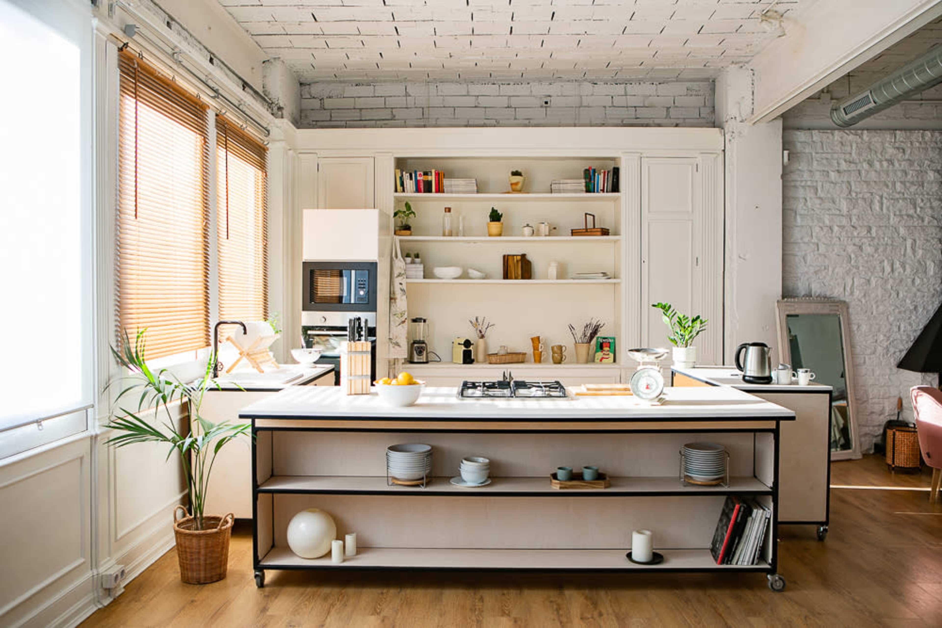 A modern kitchen features an island with a cooktop, open shelving displaying dinnerware, and large windows allowing natural light.