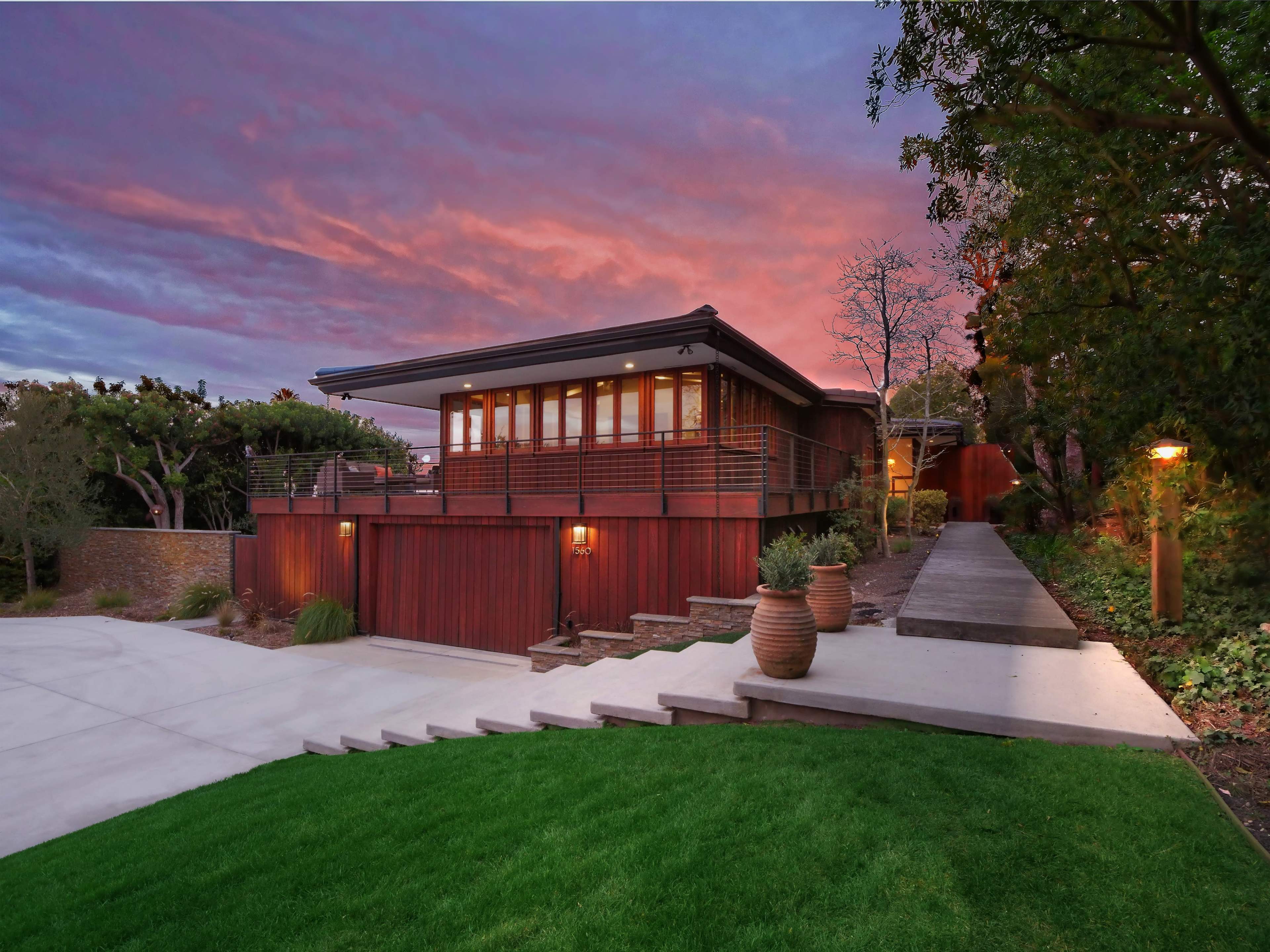 The image shows a modern house with a wooden exterior, situated on a landscaped property under a colorful sky at sunset.