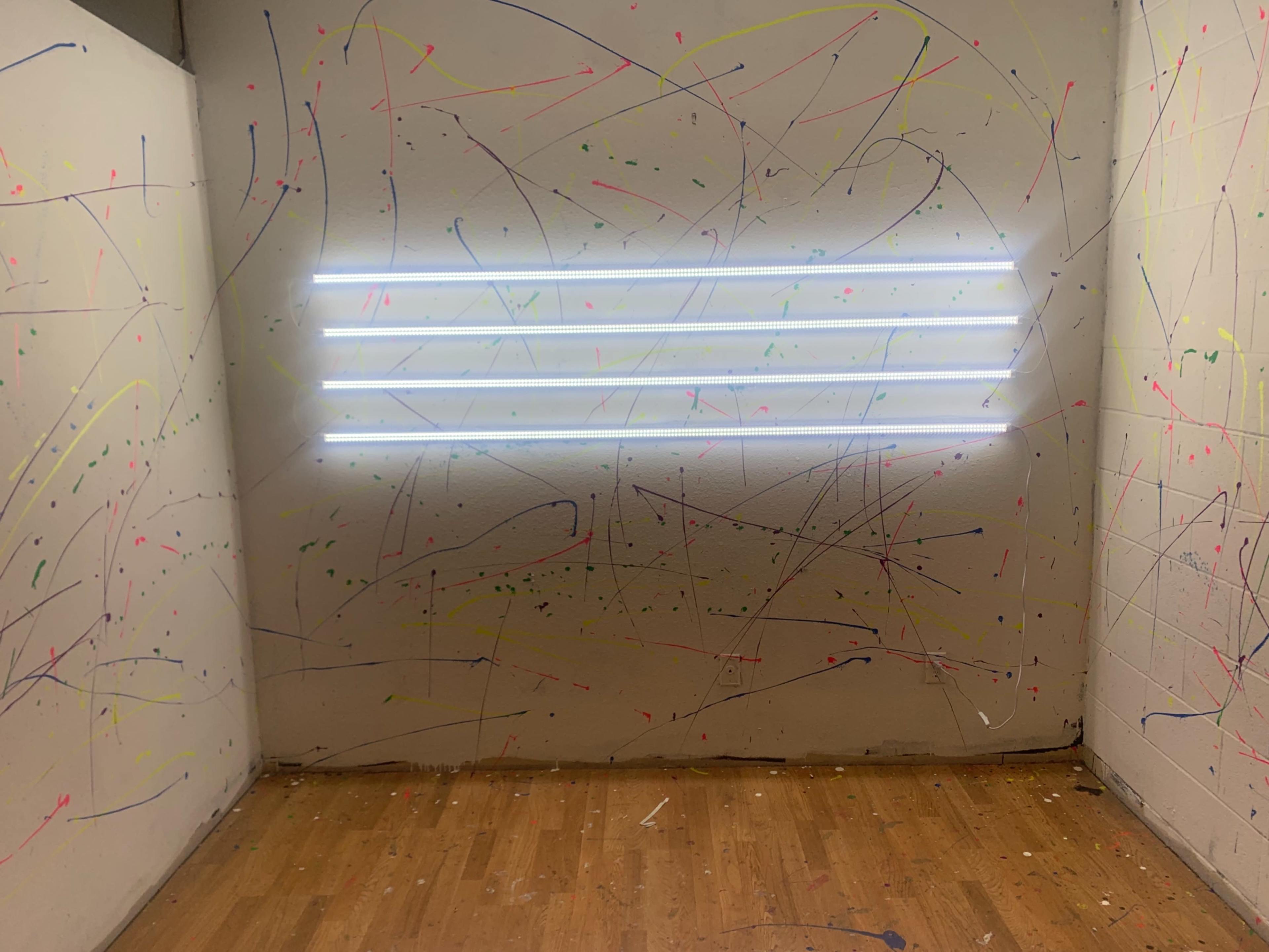 A wall with splattered paint in various colors and a set of four fluorescent light strips mounted horizontally on the surface.
