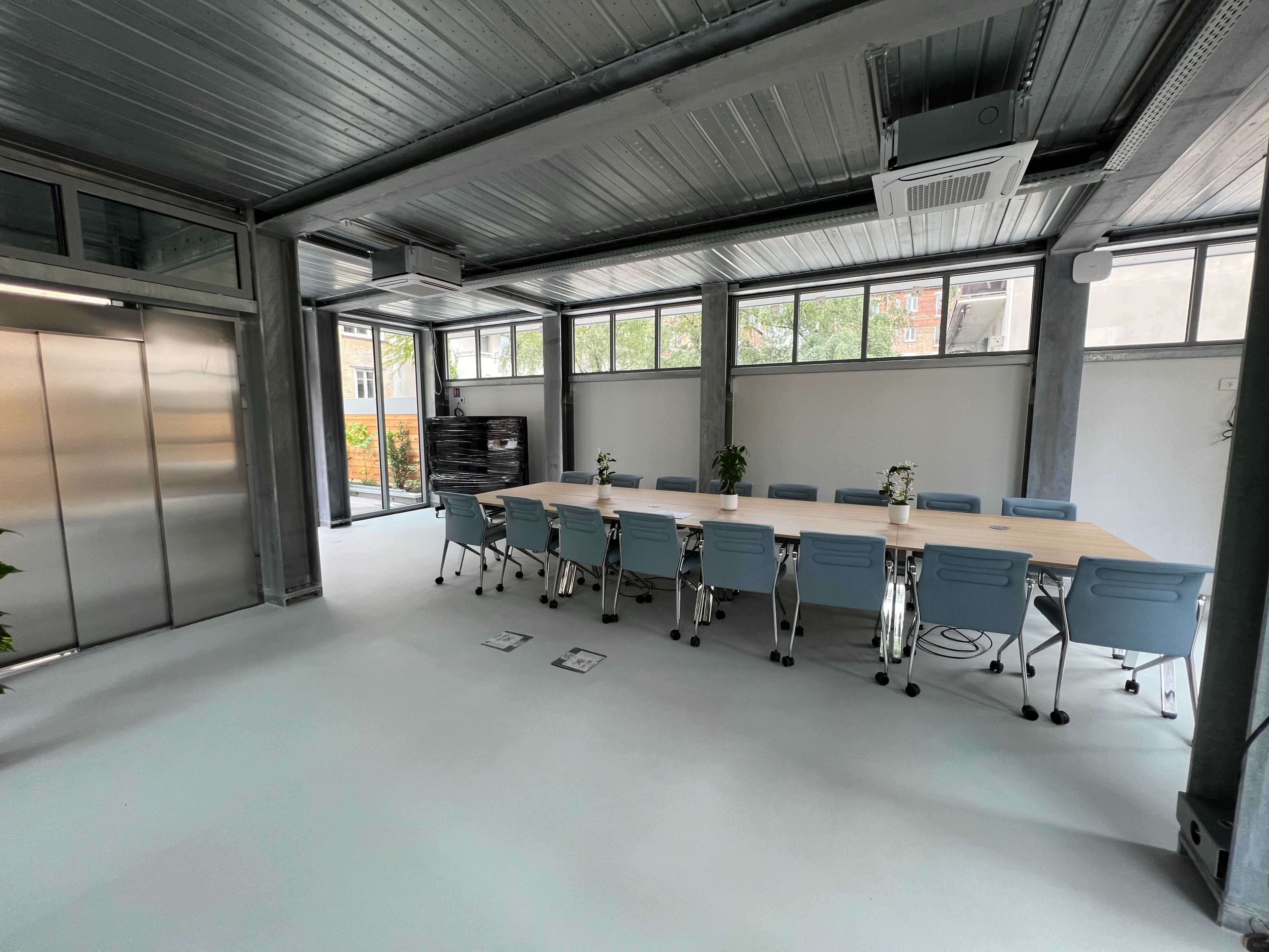 A modern meeting room features a long wooden table surrounded by blue chairs, with large windows and an elevator in the background.
