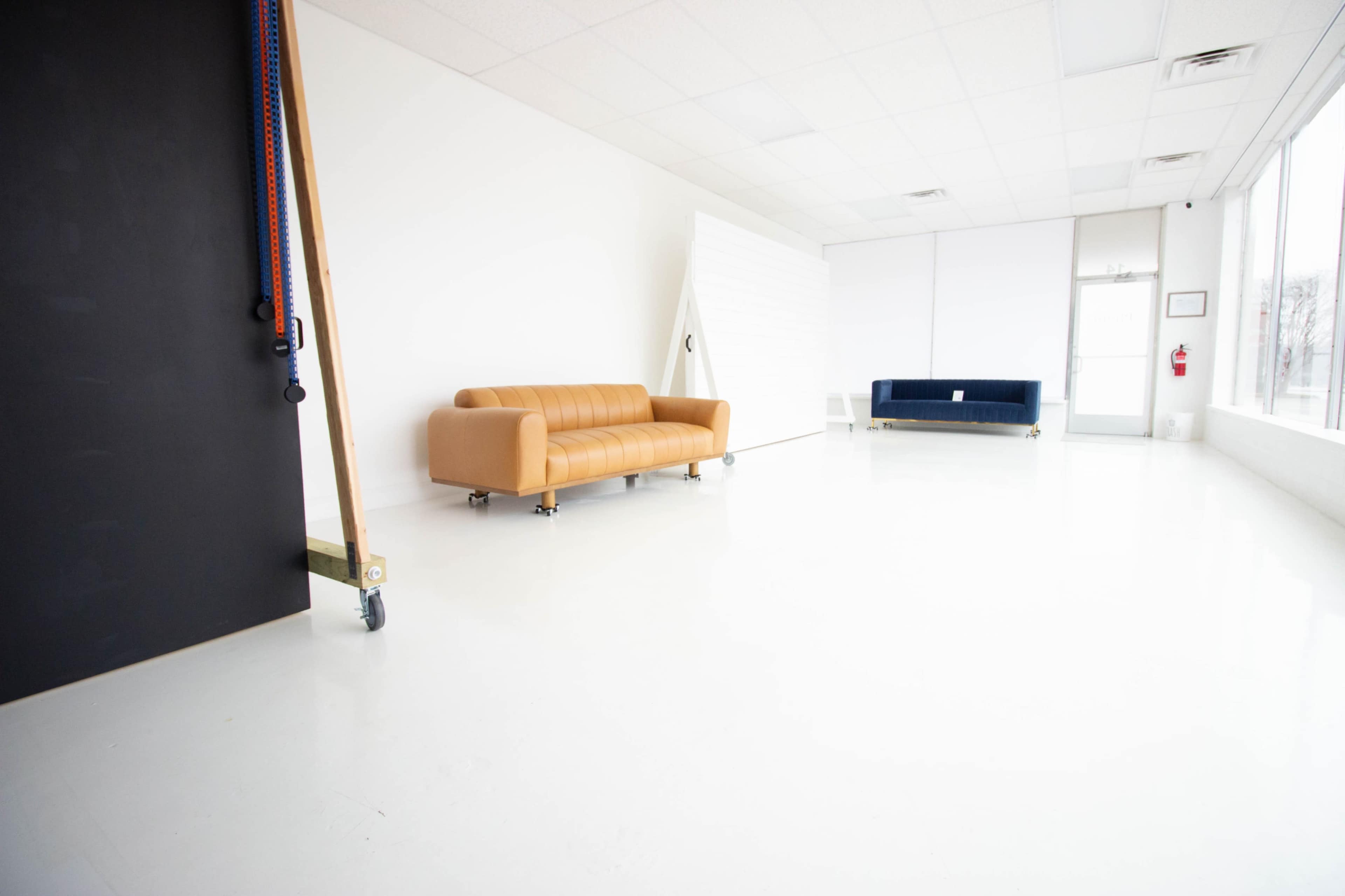 The image shows a spacious, empty room with two sofas, one brown leather and one dark blue, and a large window with natural light.
