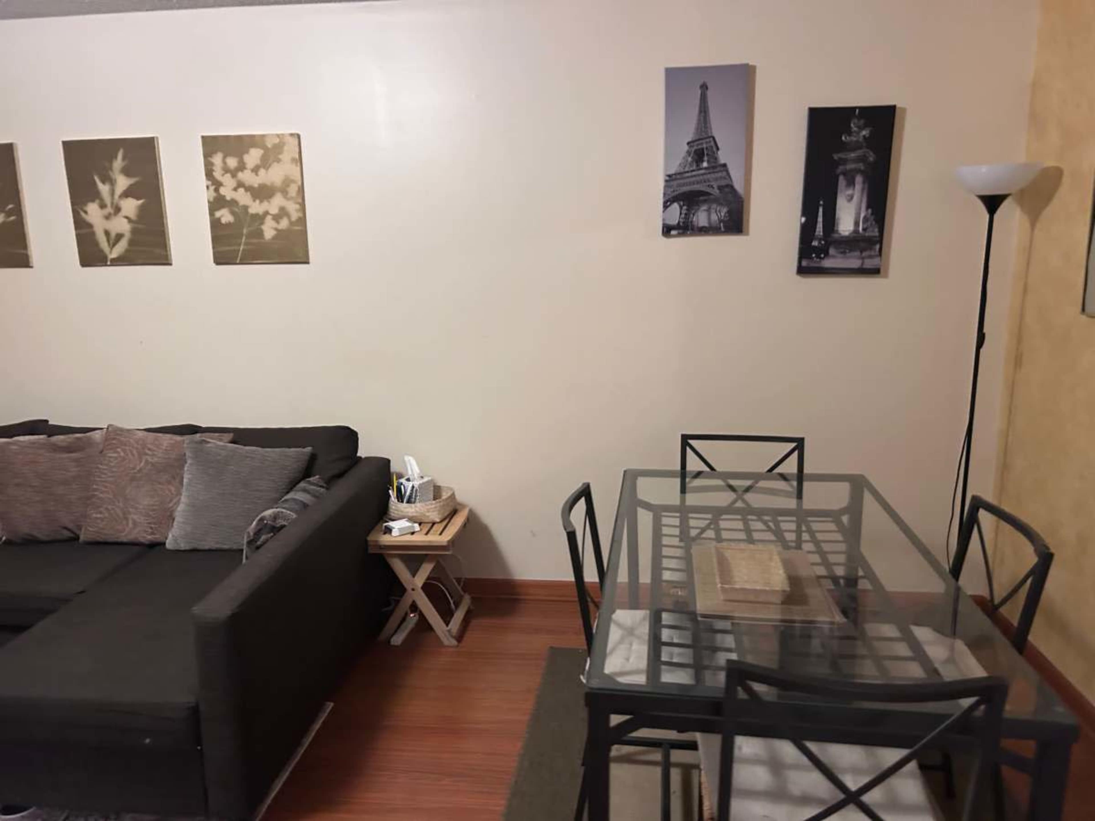 The image shows a living room with a black sofa, a small side table, a dining table with chairs, and framed artwork on the walls.