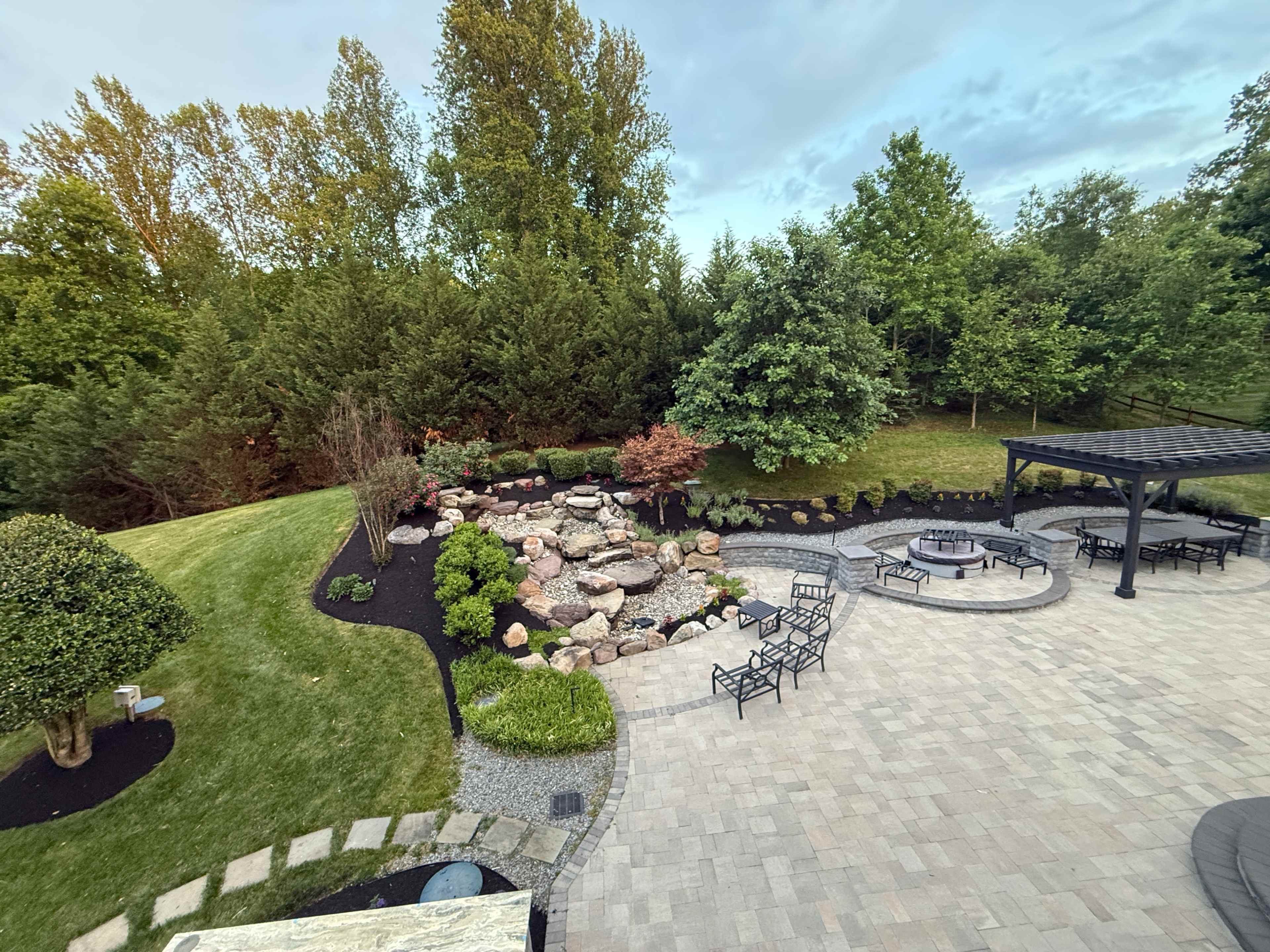 The image shows a landscaped backyard featuring a stone pond surrounded by rocks and plants, with seating areas arranged on a paved patio.