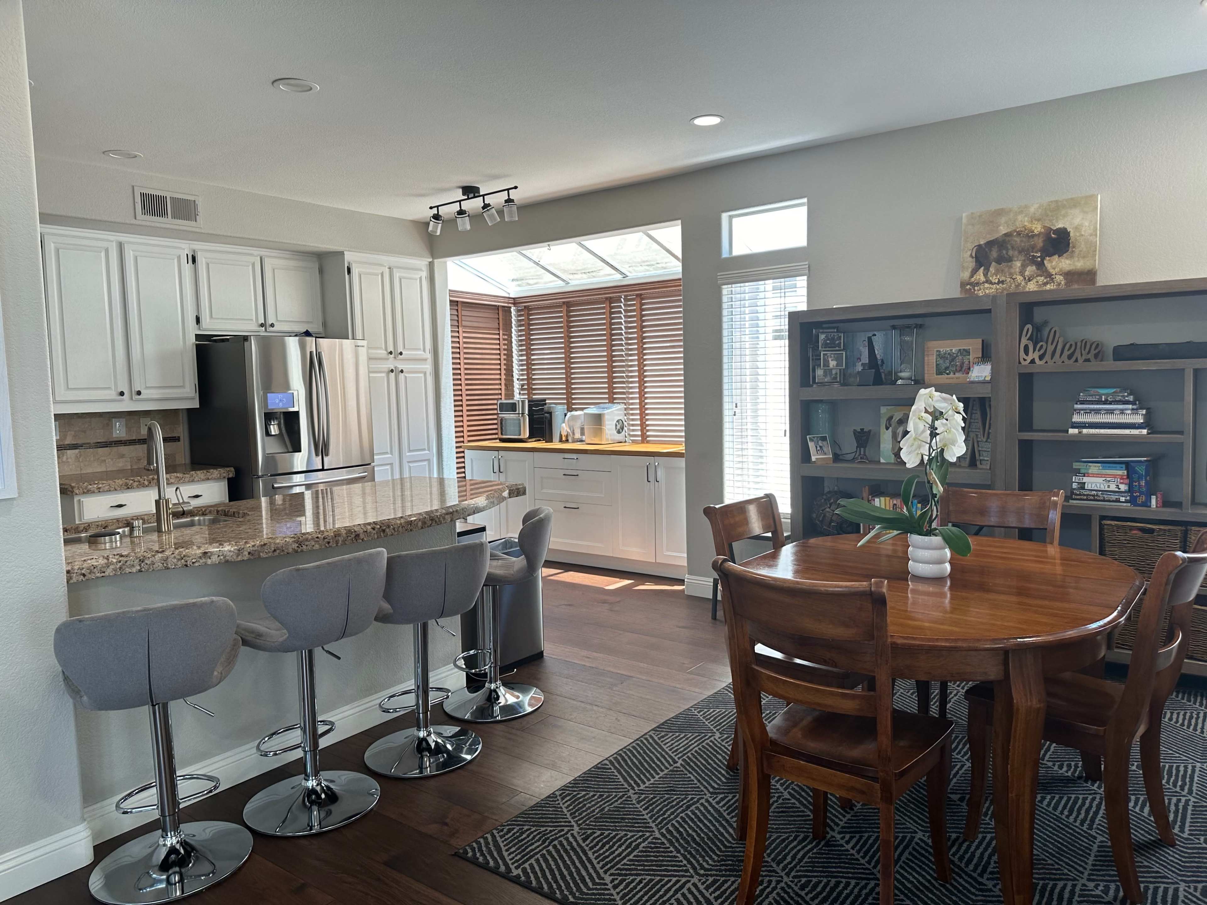A modern kitchen and dining area featuring a bar with four high stools, a wooden dining table with six chairs, and large windows with wooden blinds allowing natural light to enter.