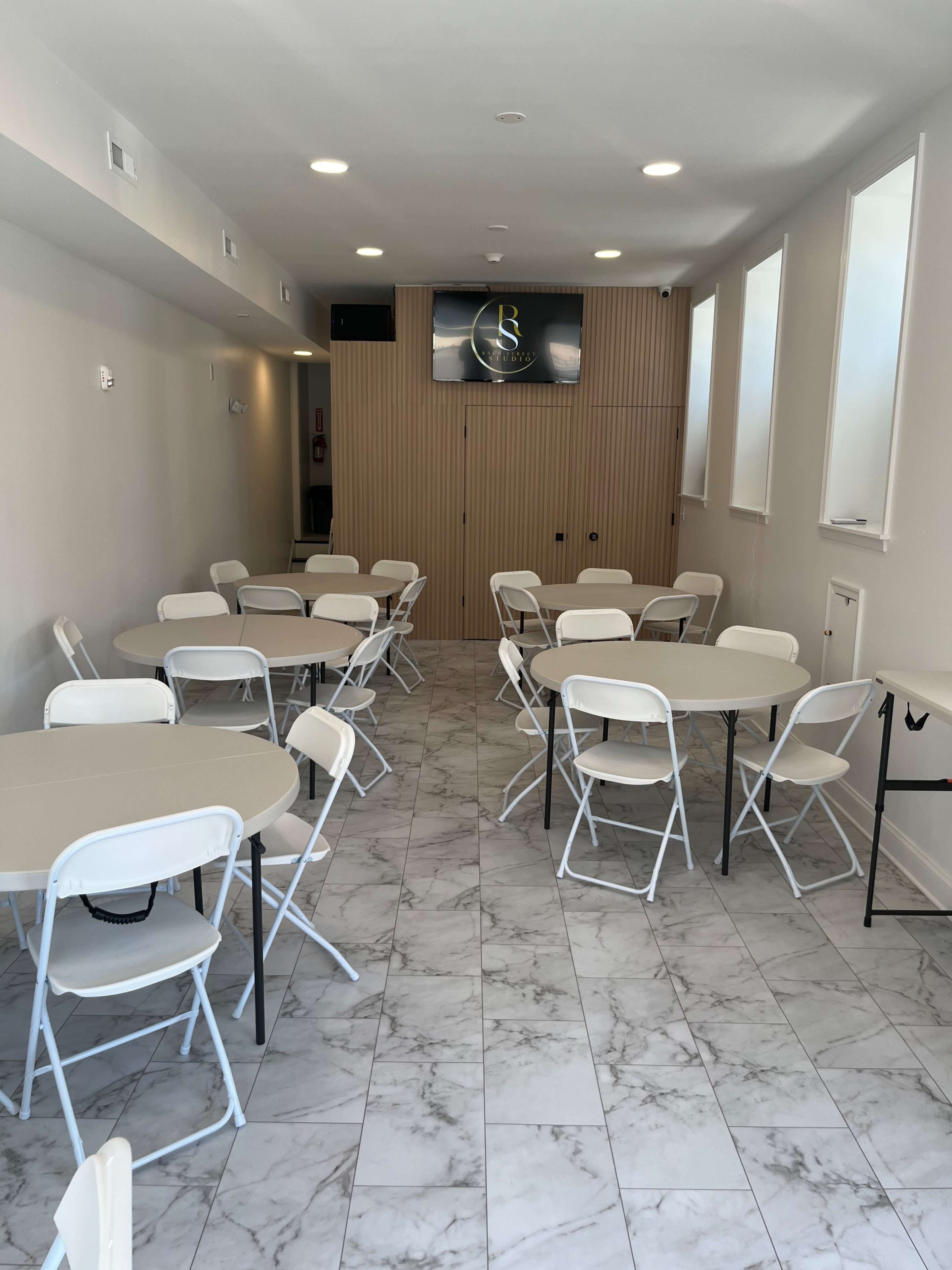 The image shows an empty, well-lit room with several round tables and chairs arranged neatly on a marble floor, featuring a wall-mounted screen.