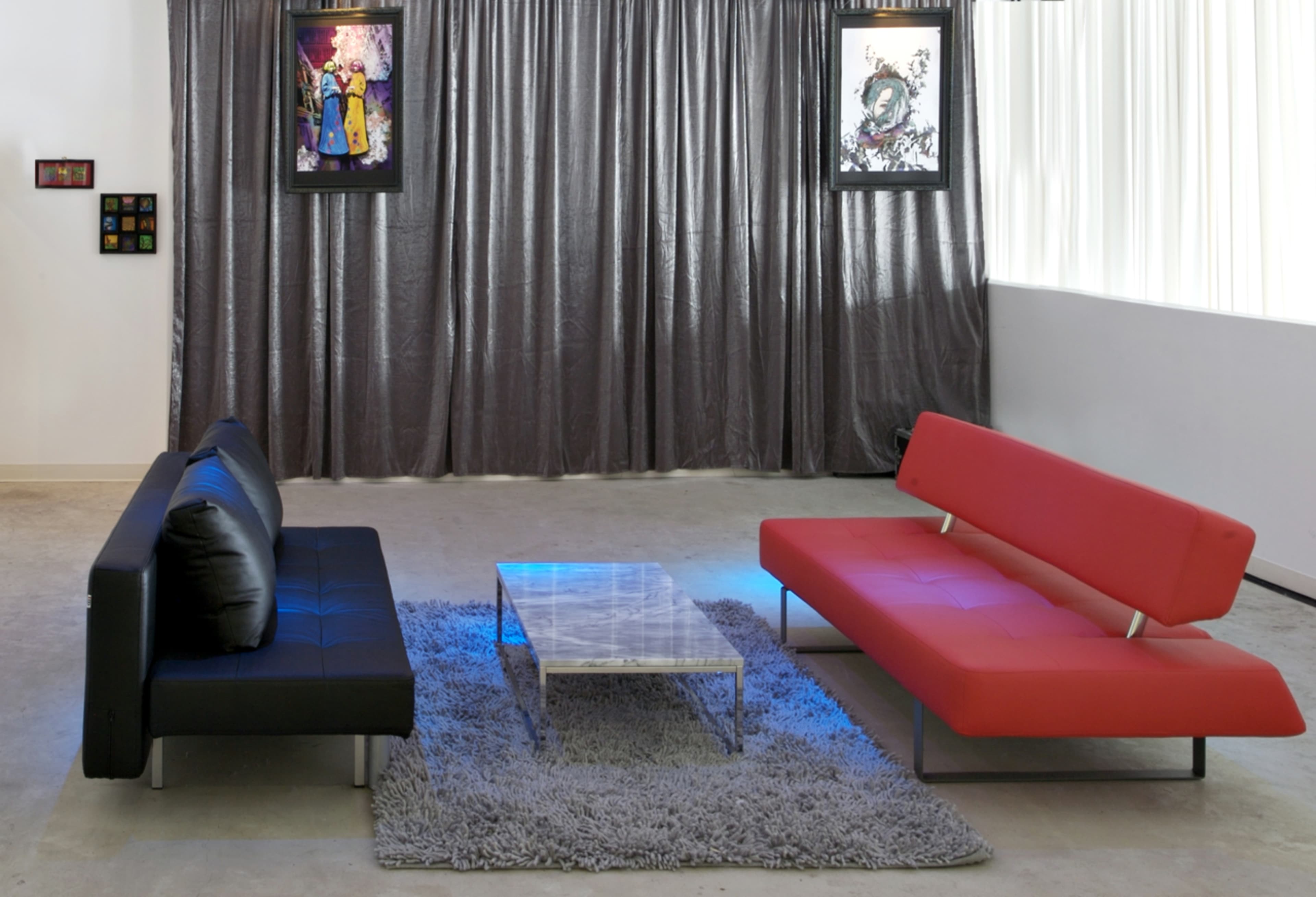 The image shows a modern living room with a black sofa and a red sofa facing each other over a glass coffee table, set against a backdrop of gray curtains and framed artwork on the walls.