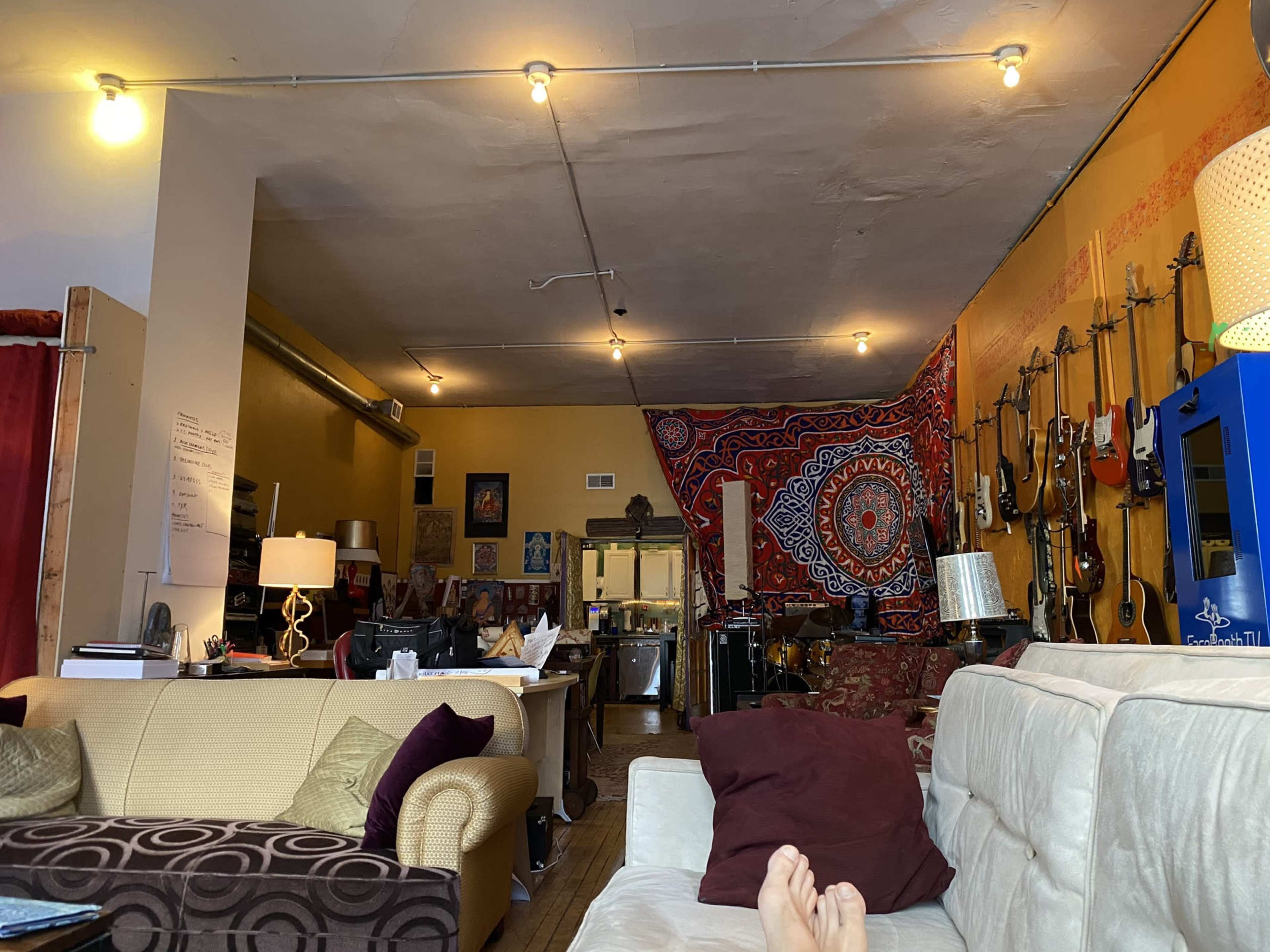 A cozy living space with seating, a colorful wall hanging, guitars displayed on one side, and a work area visible in the background.