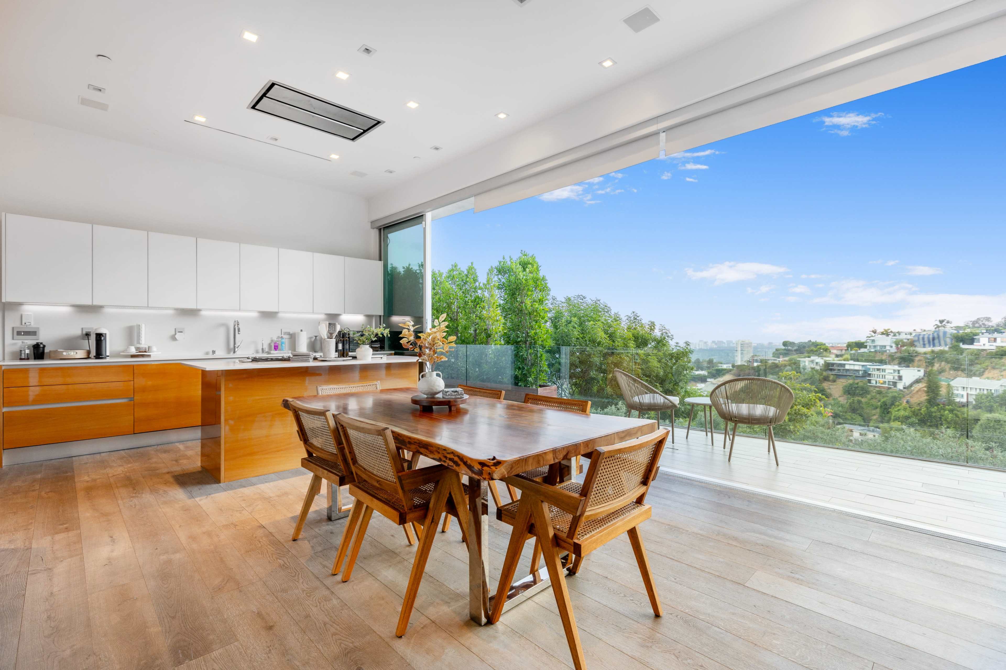 A modern kitchen with wooden cabinets and a dining area featuring a large wooden table, overlooking a scenic view through floor-to-ceiling glass doors.