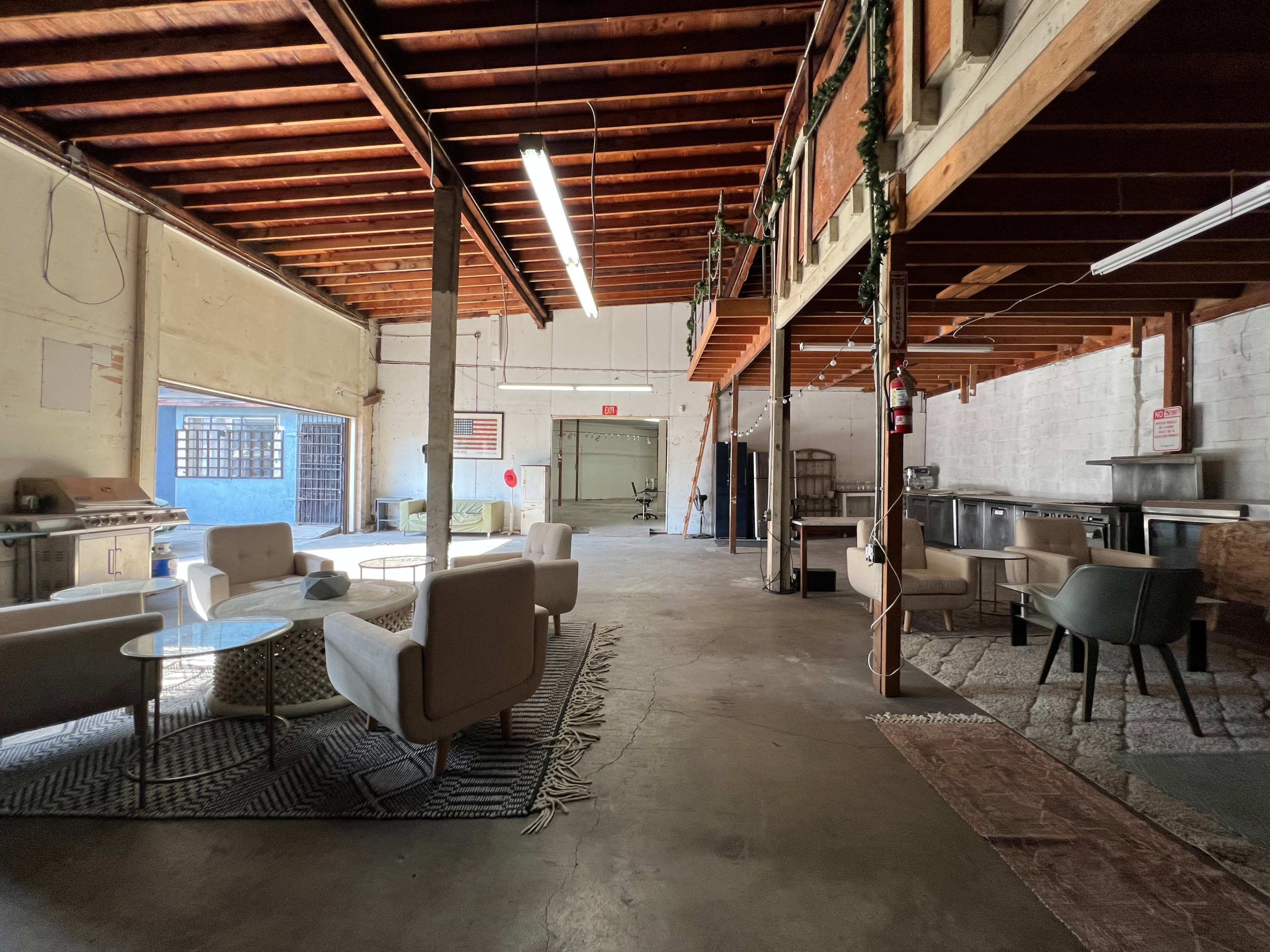 An open warehouse space with a seating area featuring modern chairs and tables, illuminated by overhead lights.