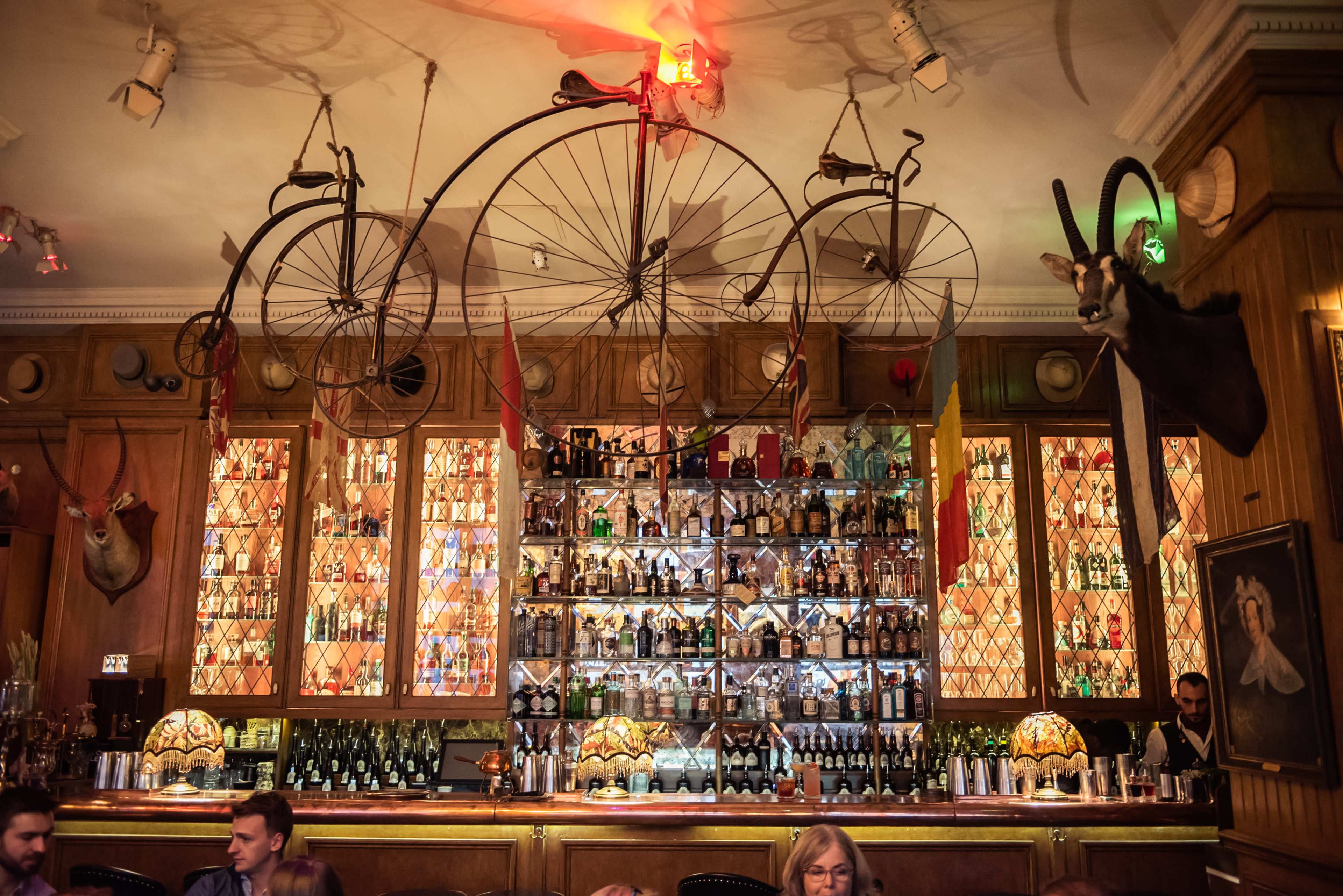 The interior of a bar features an extensive collection of bottles displayed behind the counter, with antique bicycles and mounted animal heads decoratively arranged above.