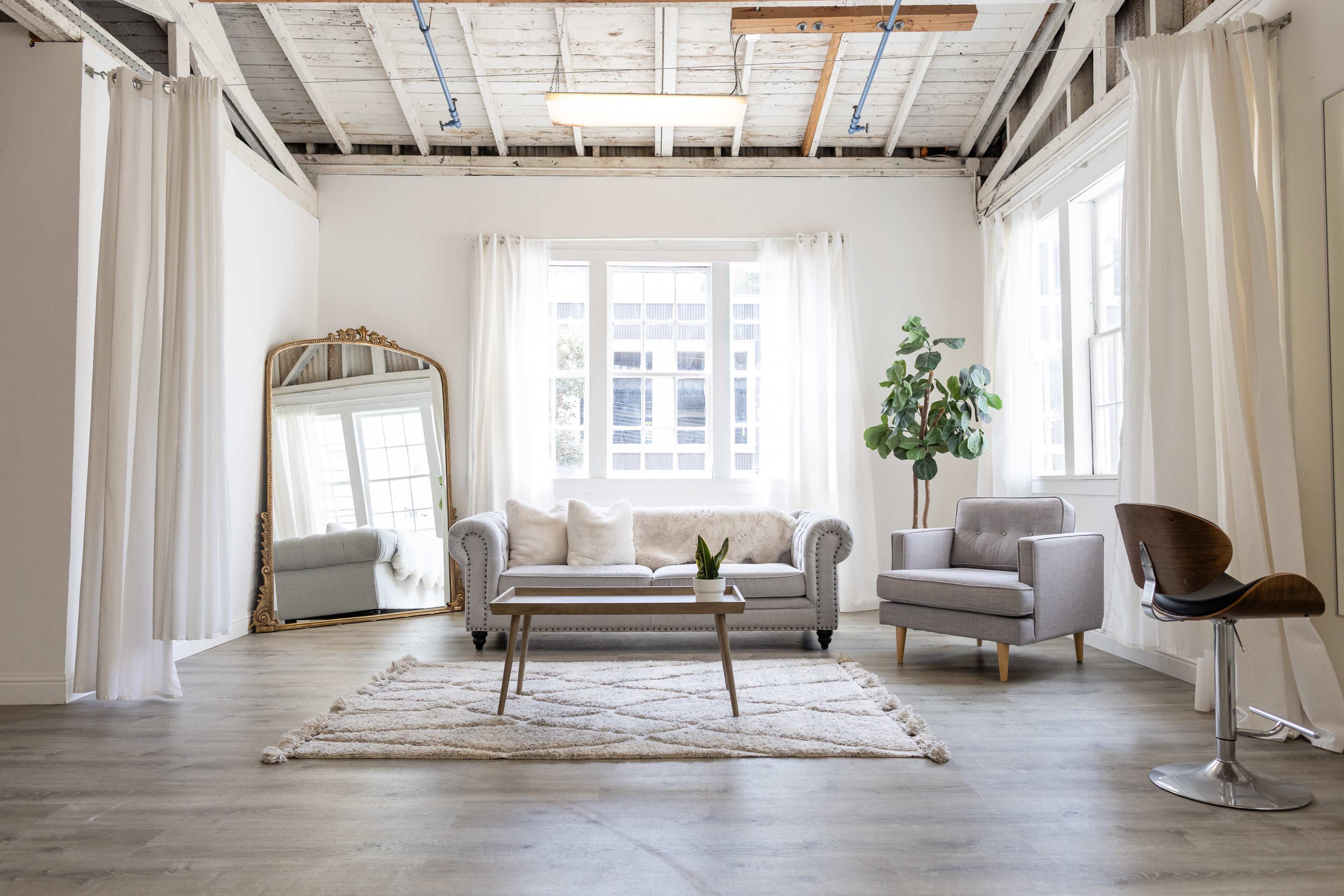 A bright, minimalist living space featuring a light-colored couch, a small coffee table, an armchair, and a large mirror, all set on wooden flooring with a rug in the center.