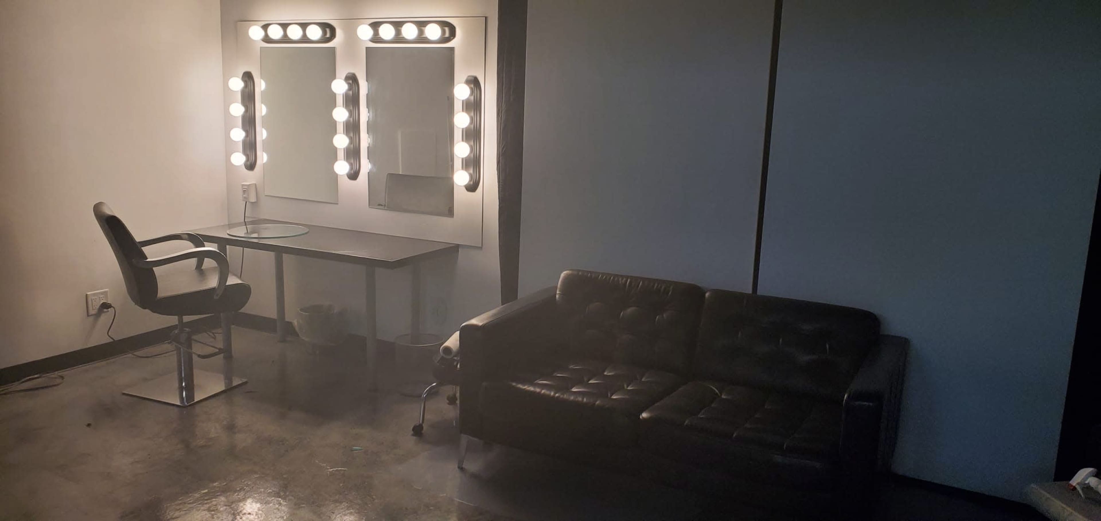The image shows a room with a black leather couch, a mirrored vanity with light bulbs, and a desk chair, all set against a simple backdrop.