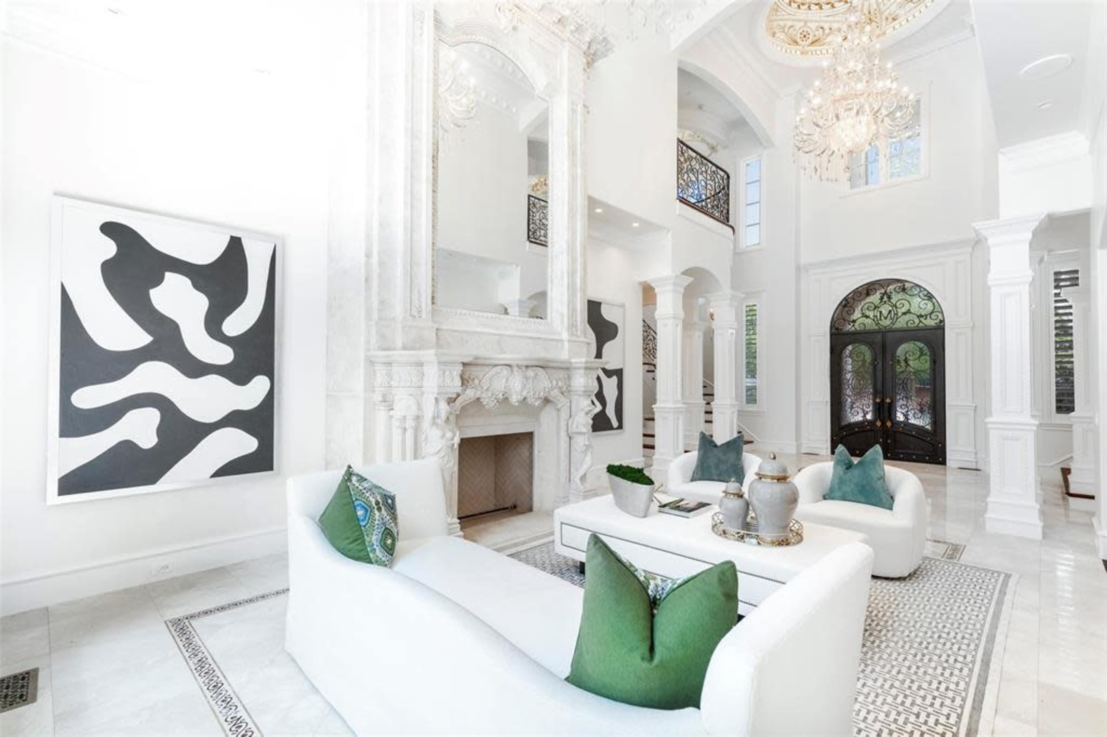 The image features a spacious, elegantly designed living room with a large marble fireplace, high ceilings, and contemporary art pieces on the walls.
