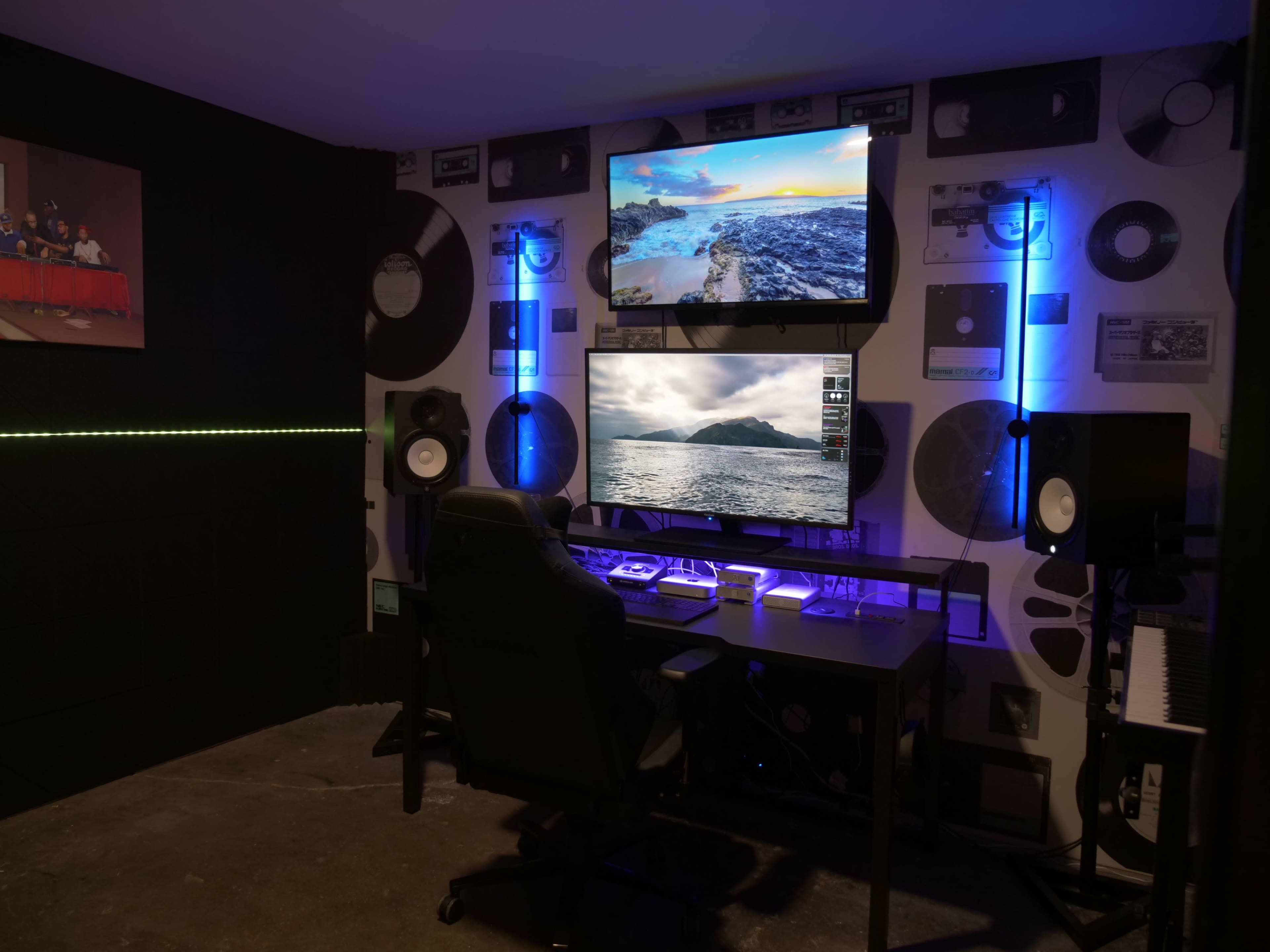 A modern workspace features a large desk with dual monitors displaying landscapes, surrounded by vinyl records on the wall and ambient lighting.